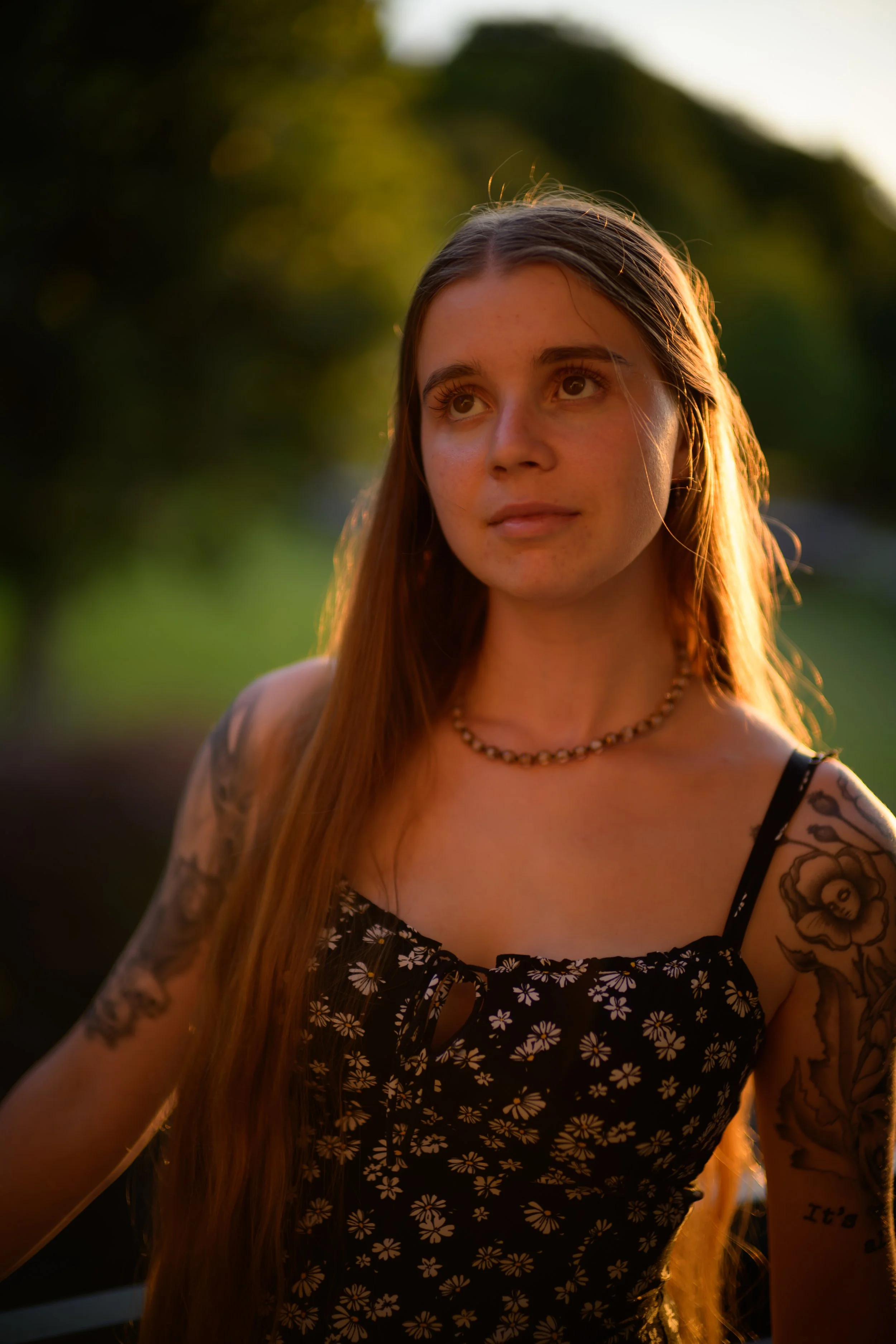 Junge Frau mit Tattoos und langen Haaren, trägt ein Sommerkleid mit Blumenmuster, in einem Sonnenuntergang im Freien.