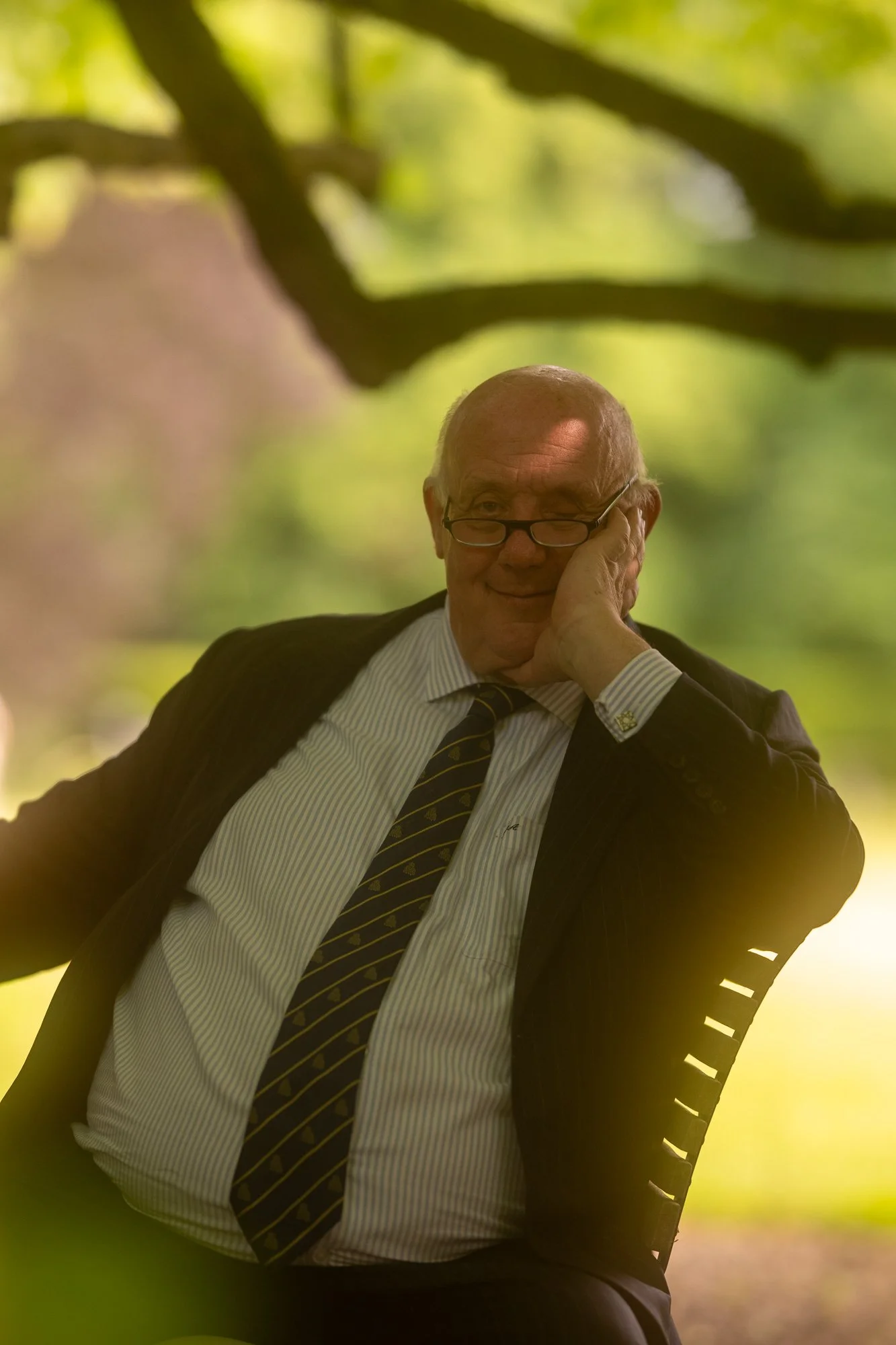 Ein älterer Mann in Anzug und Krawatte sitzt im Freien auf einem Gitterstuhl, lächelnd und die Hand an die Wange gestützt, im Hintergrund grüner Wald.