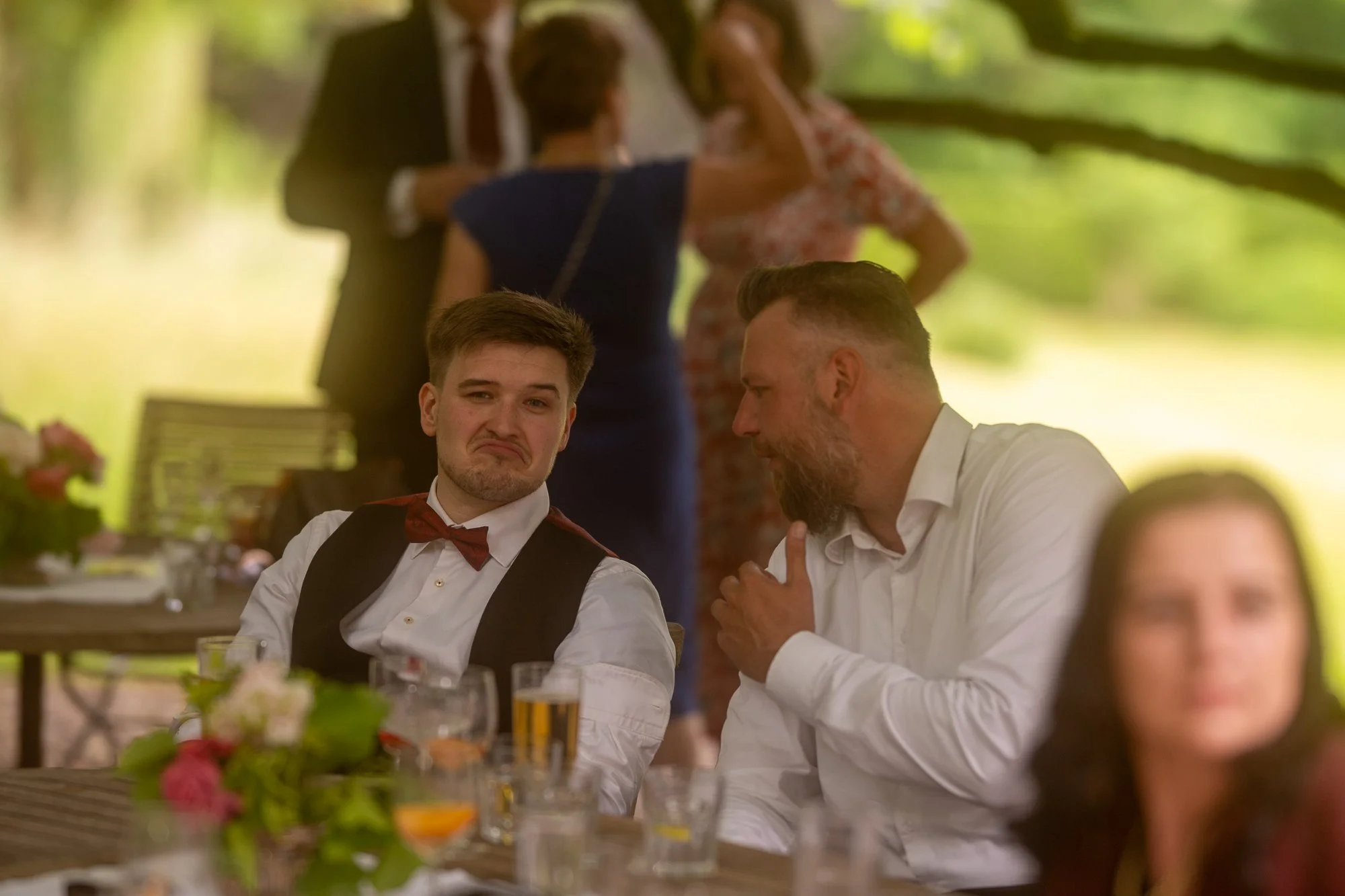 Menschen, die bei einer Feier im Freien sitzen, trinken und sich unterhalten, mit Hintergrund von Bäumen und Sonnenlicht.