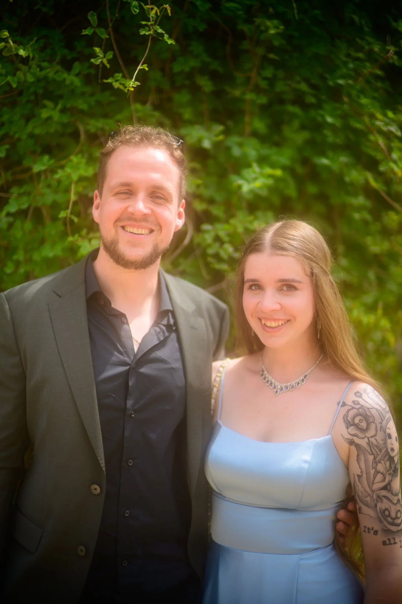 Zwei Menschen, ein Mann in einem schwarzen Anzug und eine Frau in einem weißen Kleid, stehen vor einer grünen Laubwand und lächeln in die Kamera.