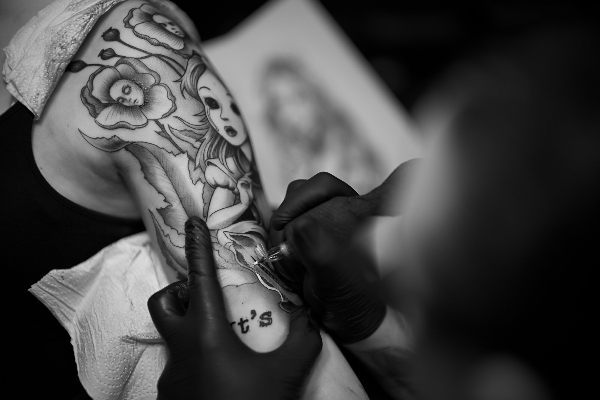 Schwarze Tätowierer arbeitet an einem Tattoo eines Mädchens mit großen Augen, umgeben von Blumen, auf dem Oberschenkel.