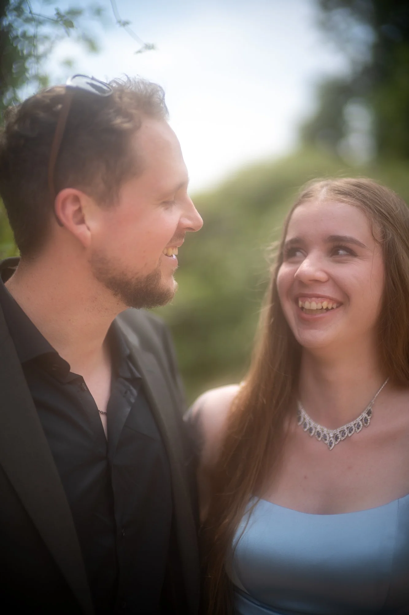 Ein Mann und eine Frau lachen sich im Freien an. Der Mann trägt eine schwarze Jacke und hat braune, kurze Haare. Das Mädchen trägt ein hellblaues Kleid und eine silberne Halskette. Im Hintergrund sind Bäume und Himmel sichtbar.