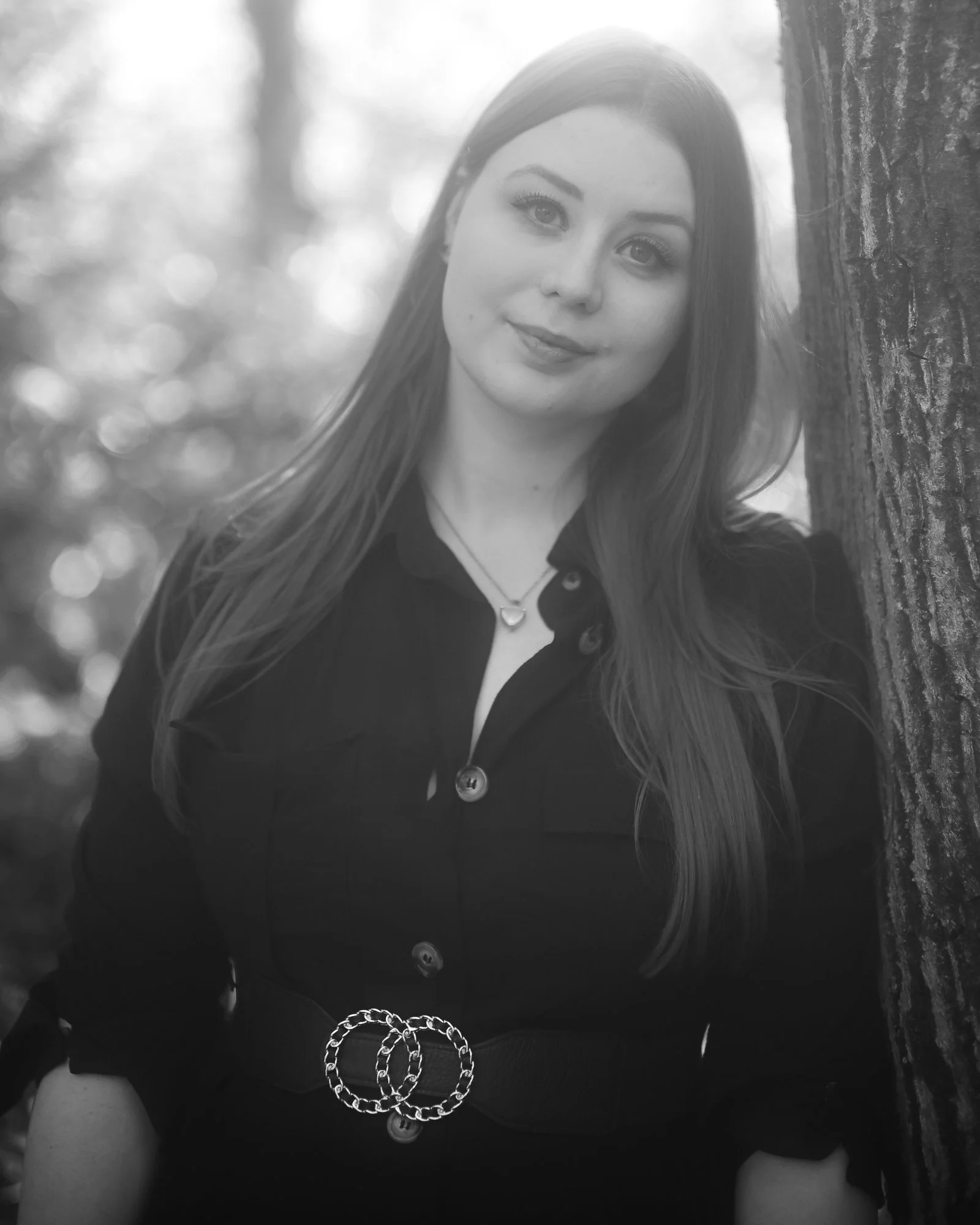 Schwarz-Weiß-Foto eines jungen Mädchens, das an einem Baum lehnt, mit langen Haaren, einem schwarzen Kleid und einer silbernen Kette, im Hintergrund unscharfe Bäume und Blätter.