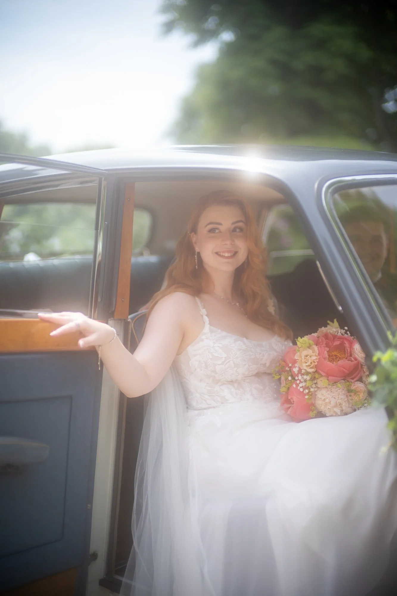 Braut in Hochzeitskleid mit Blumenstrauß im Auto bei Sonnenschein