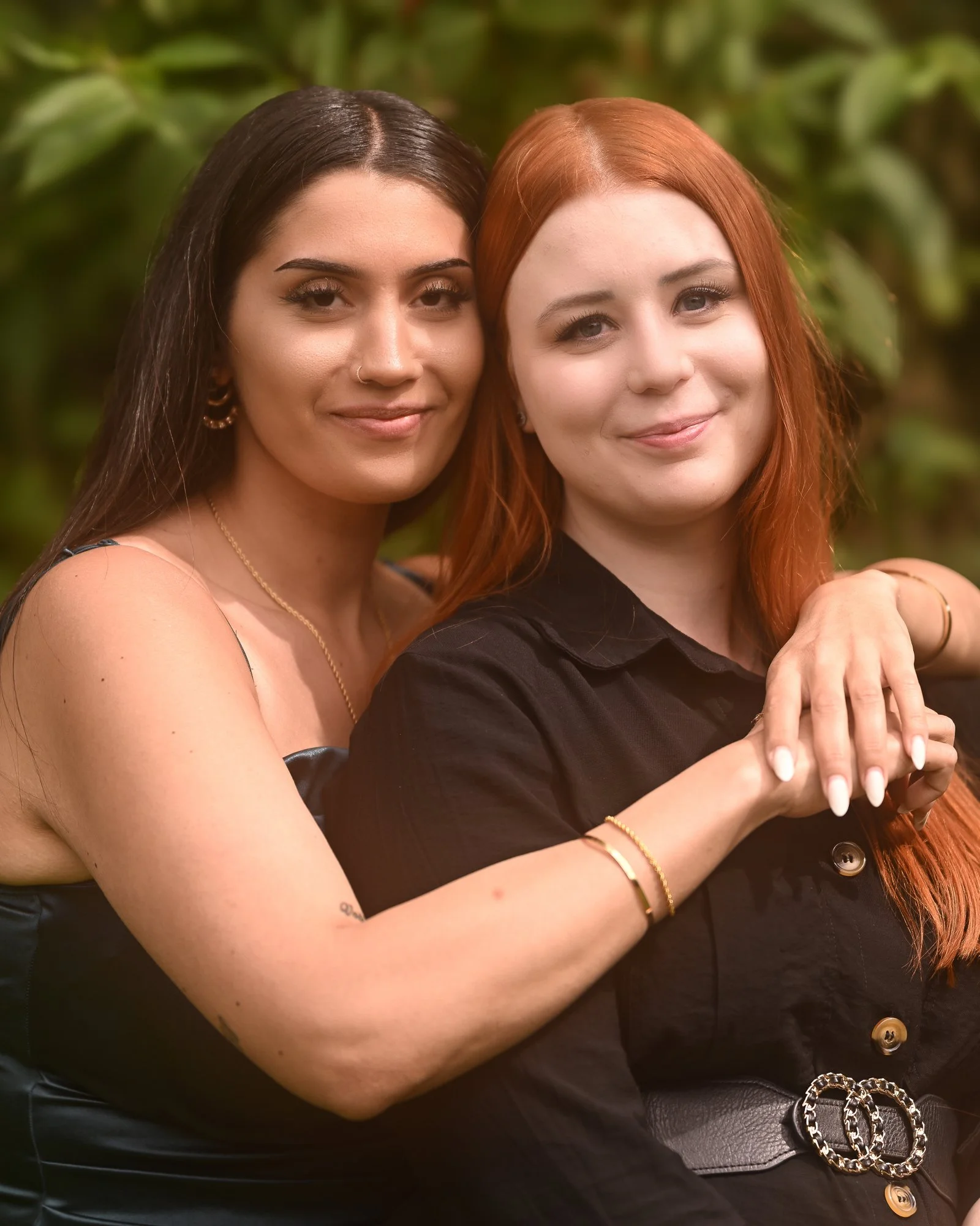 Zwei Frauen stehen eng beieinander vor einer grünen, blätterreichen Hintergrund und lächeln freundlich.