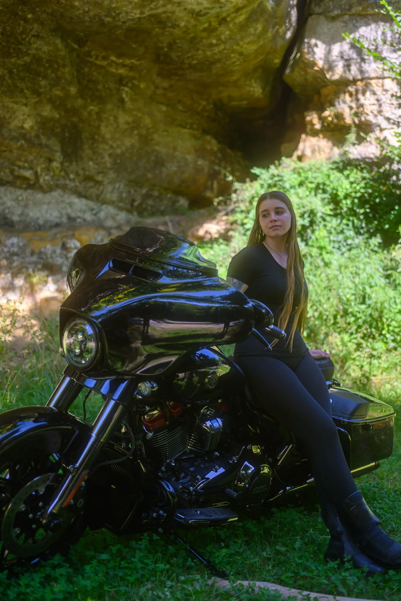 Junge Frau sitzt auf einer schwarzen Harley-Davidson-Motorrad vor felsiger, grüner Natur.