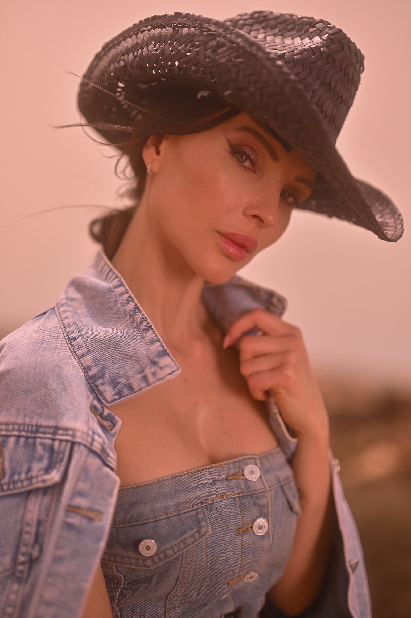 Frau trägt einen Hut und eine Jeansjacke, hält einen Lockenstab und schaut in die Kamera.