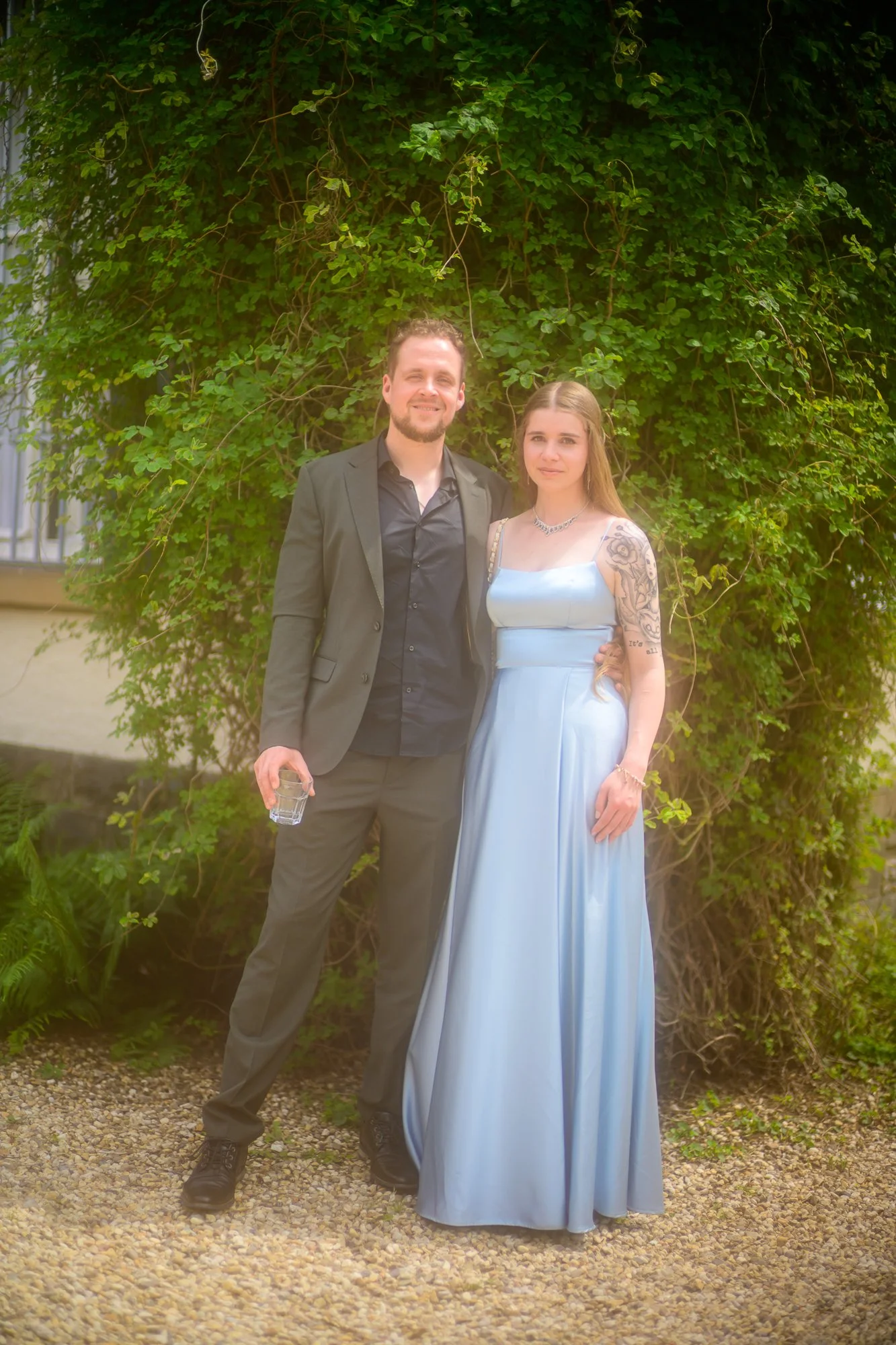 Ein Mann und eine Frau stehen nebeneinander vor einer grünen Laubwand. Der Mann trägt einen schwarzen Anzug und hält ein Trinkglas. Die Frau trägt ein langes, hellblaues Kleid und hat Tattoos an den Armen. Es ist taghell, und sie scheinen bei einem b