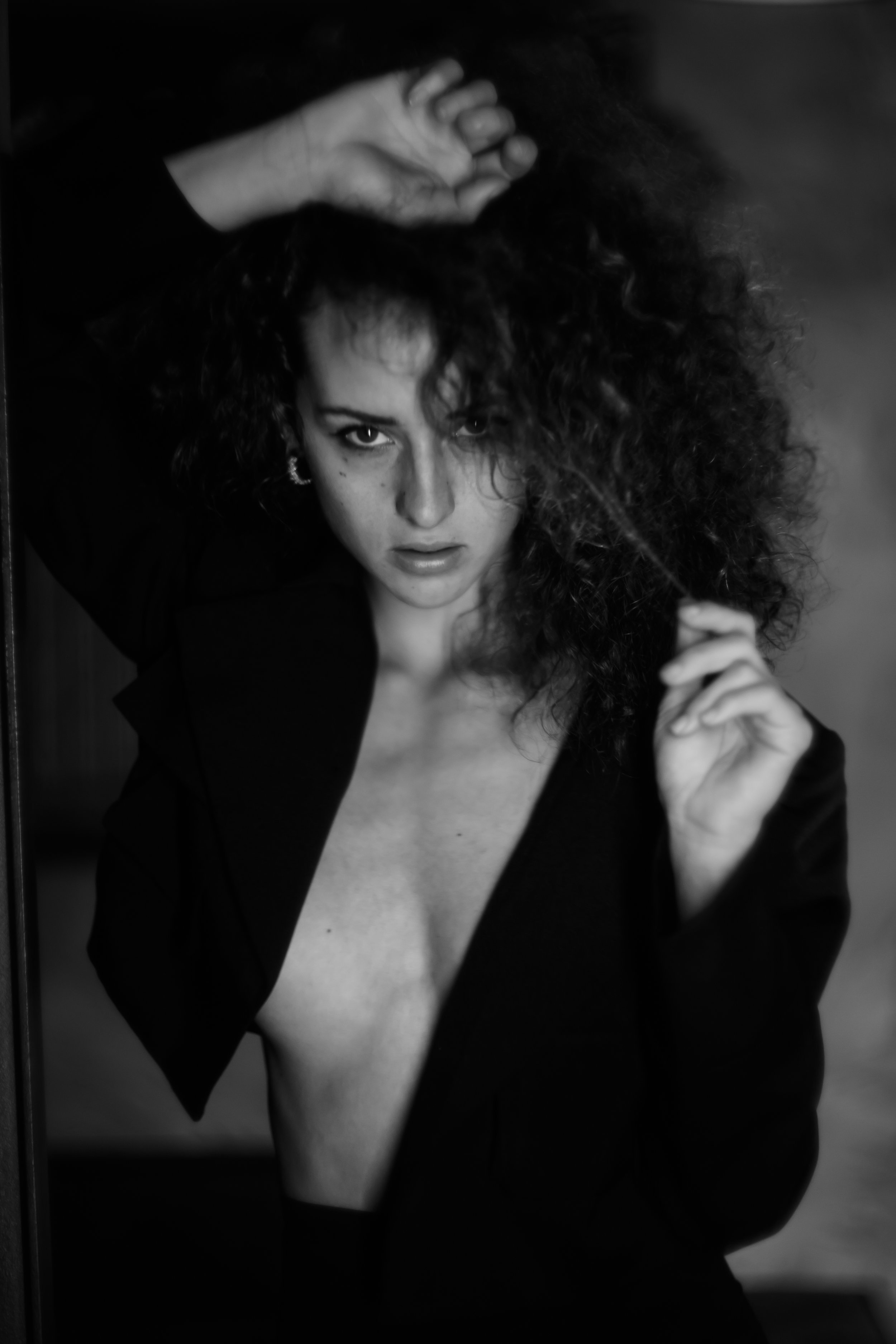 Schwarz-Weiß-Foto einer Frau mit lockigem Haar, die leicht nackt ist und einen schwarzen Blazer trägt, während sie in einen Spiegel schaut.