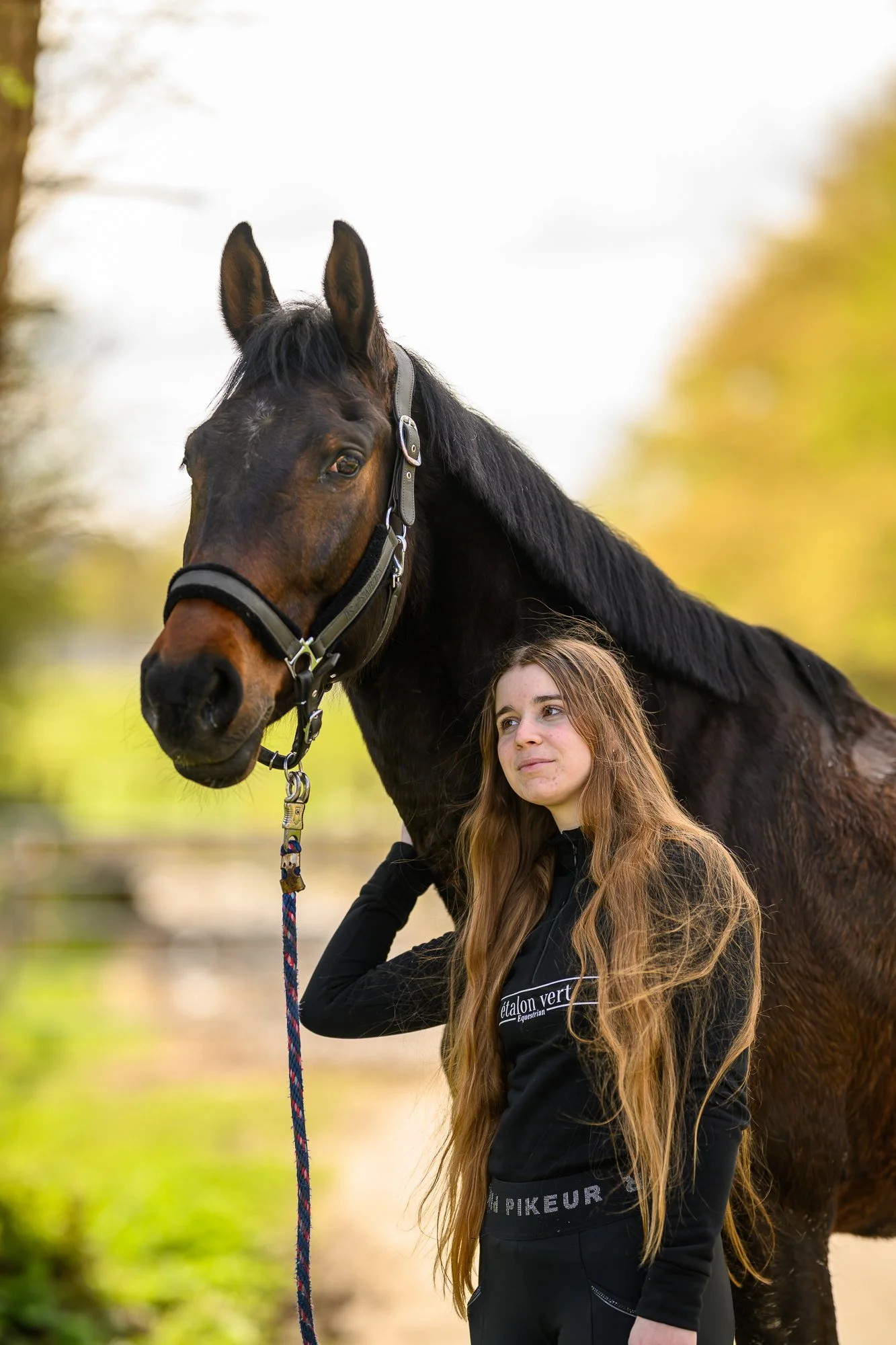 Ein junges Mädchen mit lang, lockigem braunem Haar steht neben einem schwarzen Pferd mit braunem Kopf und Hals. Das Mädchen trägt ein schwarzes Sportshirt mit weißen Aufdruck und hält die Leine des Pferdes. Die Szene ist im Freien mit unscharfem grün