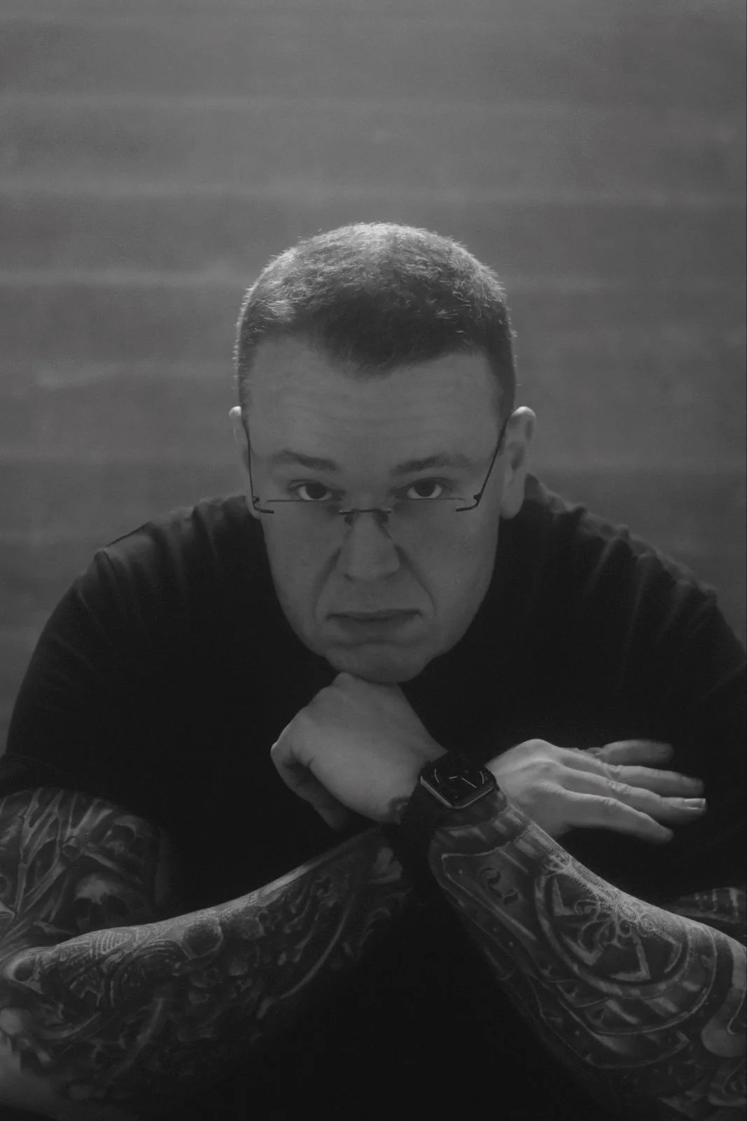 Schwarz-weiß-Foto eines Mannes mit Brille, tätowierten Armen, dunklem Haar und dunklem T-Shirt, der nach vorne schaut, mit den Armen auf einem Tisch oder einer Oberfläche ruhend.