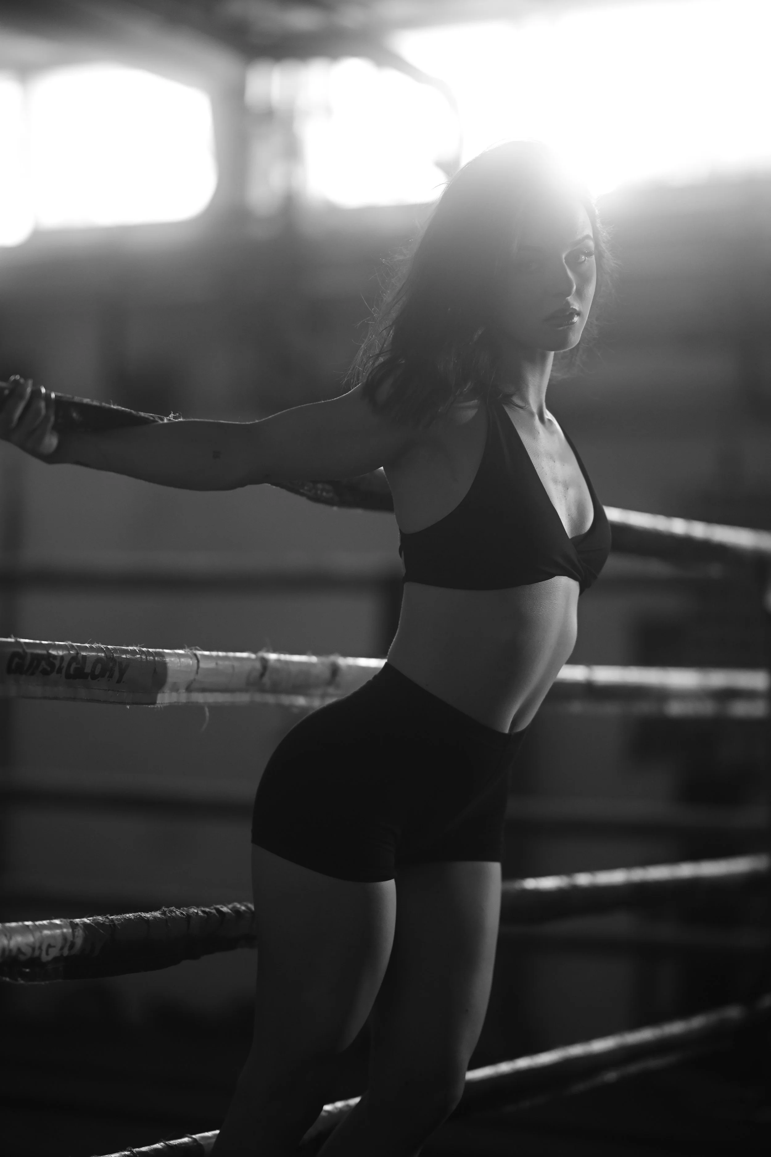 Schwarz-weiß-Foto einer jungen Frau im Fitnessstudio, die in eine Ecke ihrer Arme mit einem Seil gespannt ist. Im Hintergrund sind die Ringe eines Boxrings sichtbar. Die Frau trägt Sportbekleidung und wirkt entschlossen.