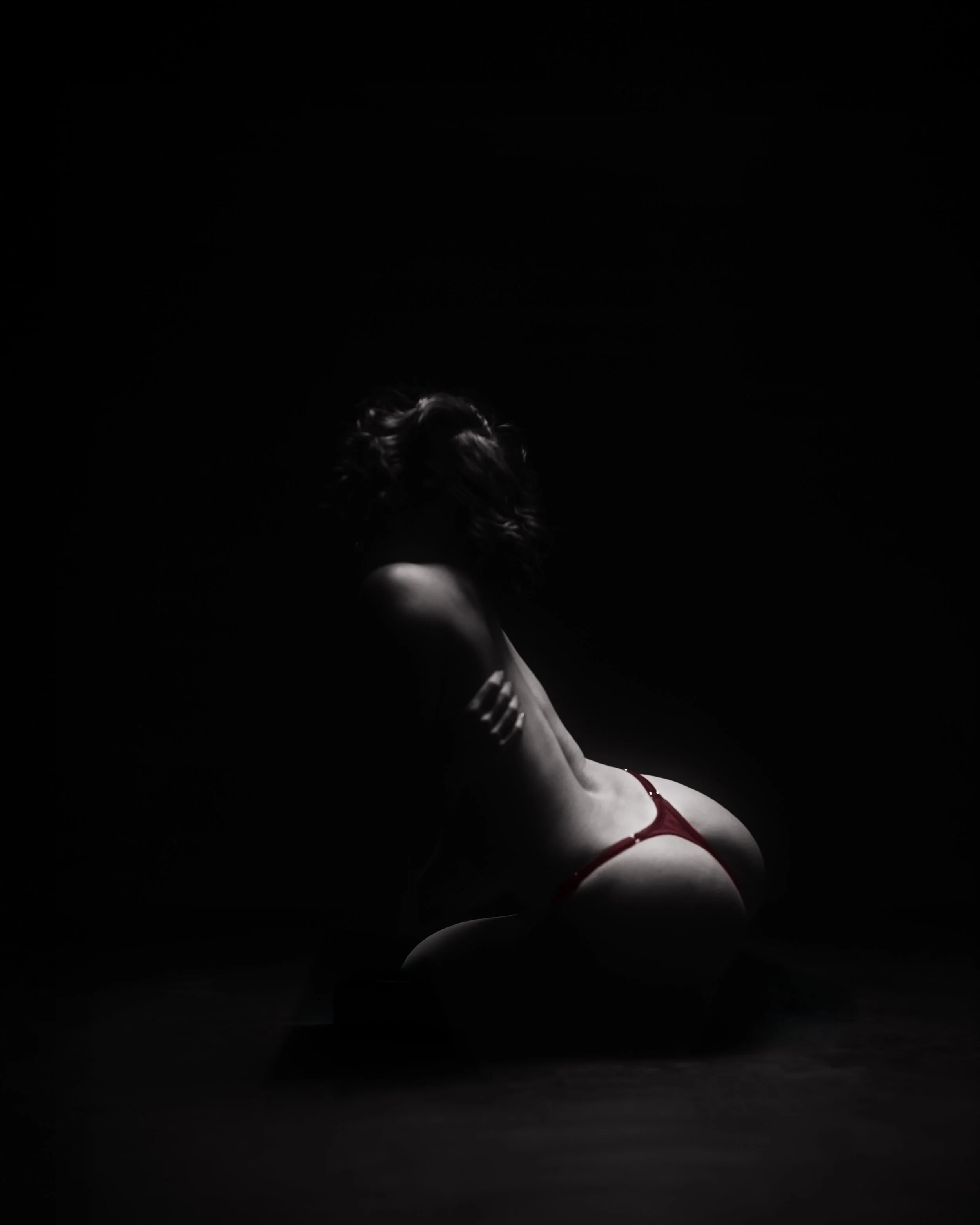 Eine Person in rotem Dessous posiert in einem dunklen Raum, das Bild ist überwiegend schwarz, mit Licht, das das Gesicht und die Schulter der Person beleuchtet.