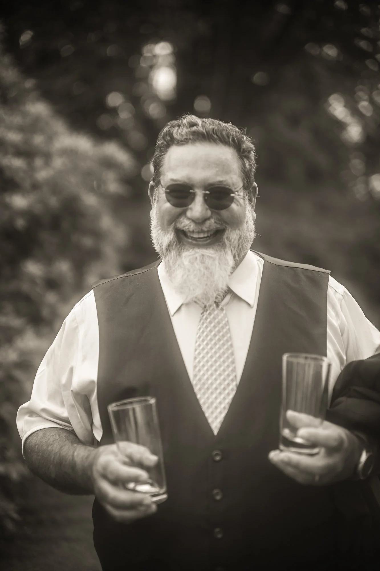 Ein fröhlicher Mann mit Brille, weißem Bart und braunen Haaren, trägt eine weiße Hemd und eine Weste, hält ein Weinglas in der Hand und lacht bei einer Feier im Freien.