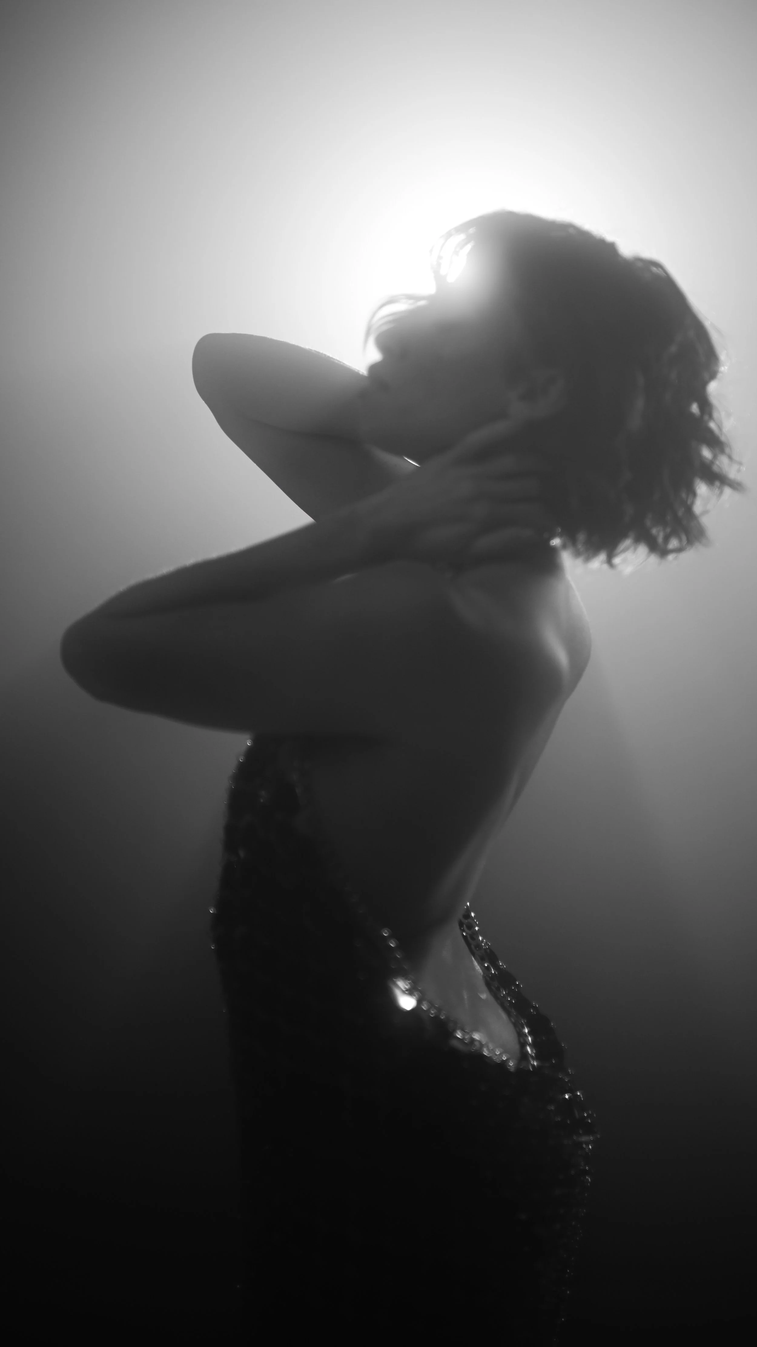 Schwarz-Weiß-Foto einer tanzenden Frau in einem eleganten Kleid, mit Haaren im Wind, hinter einer hell erleuchteten Wand.