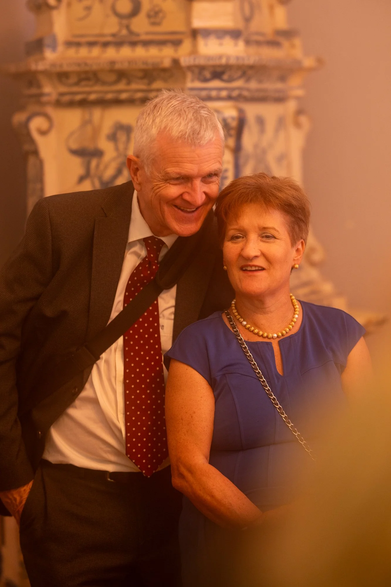 Ein älterer Mann in Anzug und Krawatte, der mit einem Lächeln eine ältere Frau in einem blauen Kleid umarmt, beide vor einer kunstvollen Wanddekoration in einem eleganten Raum.