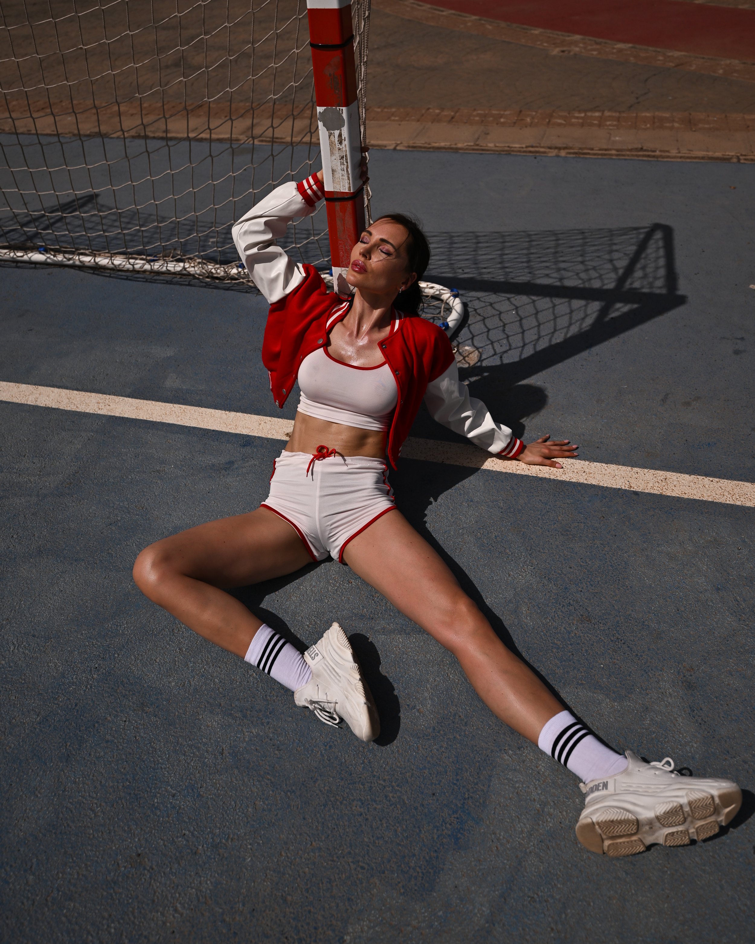 Eine Frau sitzt auf dem Boden vor einem Handballtor, trägt eine weiße Sporthose, ein rotes Jacke und weiße Turnschuhe. Sie hat die Augen geschlossen und scheint entspannt oder erschöpft zu sein.