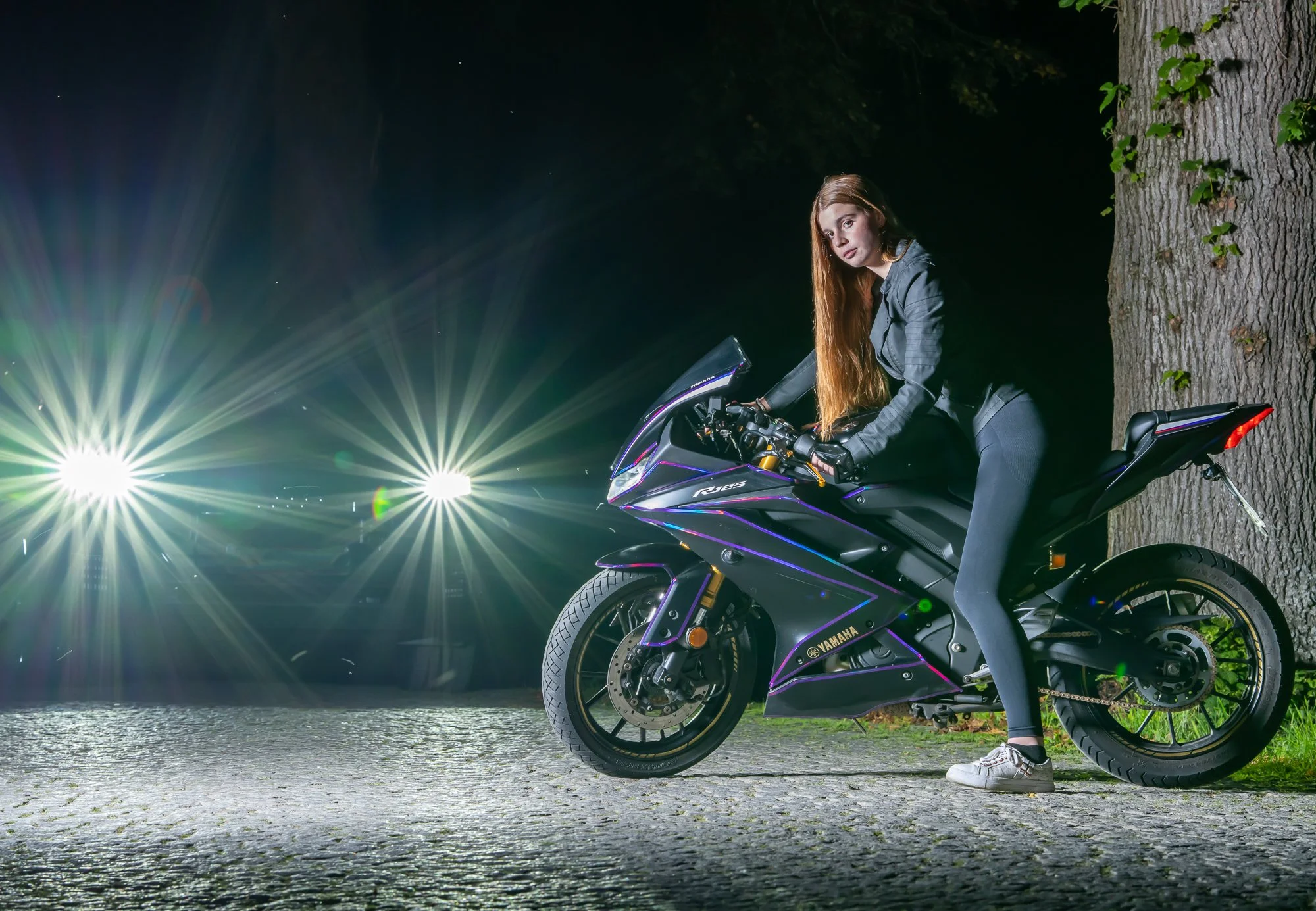 Junge Frau mit langen roten Haaren sitzt auf einer schwarzen Yamaha R125 Motorrad bei Nacht, im Hintergrund helle Straßenlaternen und bunter Himmel.