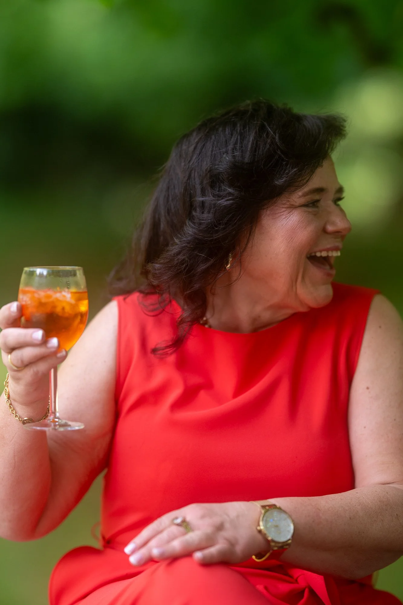 Frau in rotem Kleid hält ein Glas mit orangefarbenem Getränk, lachend, bei einer Feier im Freien.