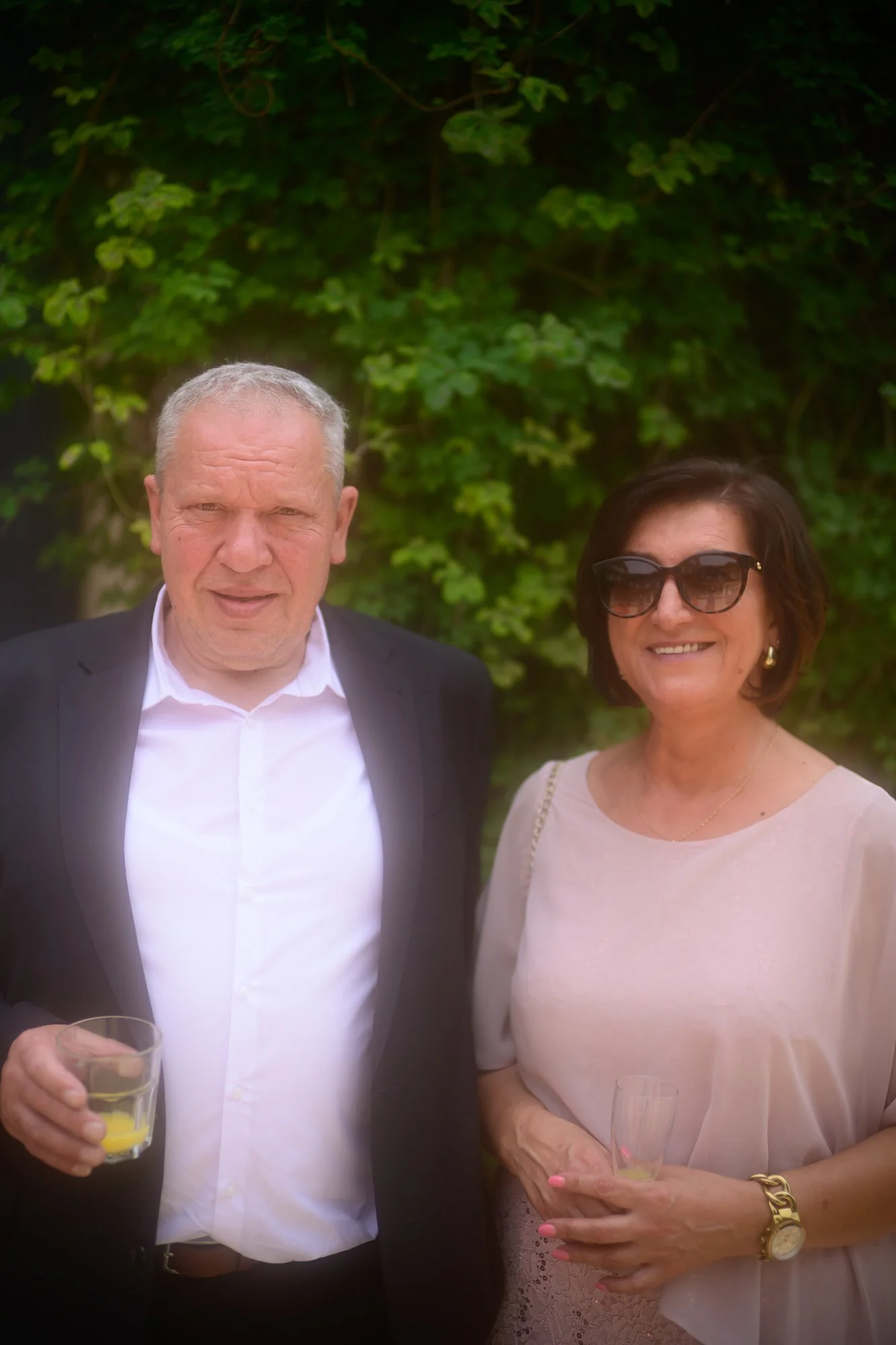 Ein Mann in einem schwarzen Anzug hält ein Glas mit Orangensaft, daneben eine Frau mit Sonnenbrille in einem hellen Kleid, beide bei einem Anlass im Freien vor grünem Laub.