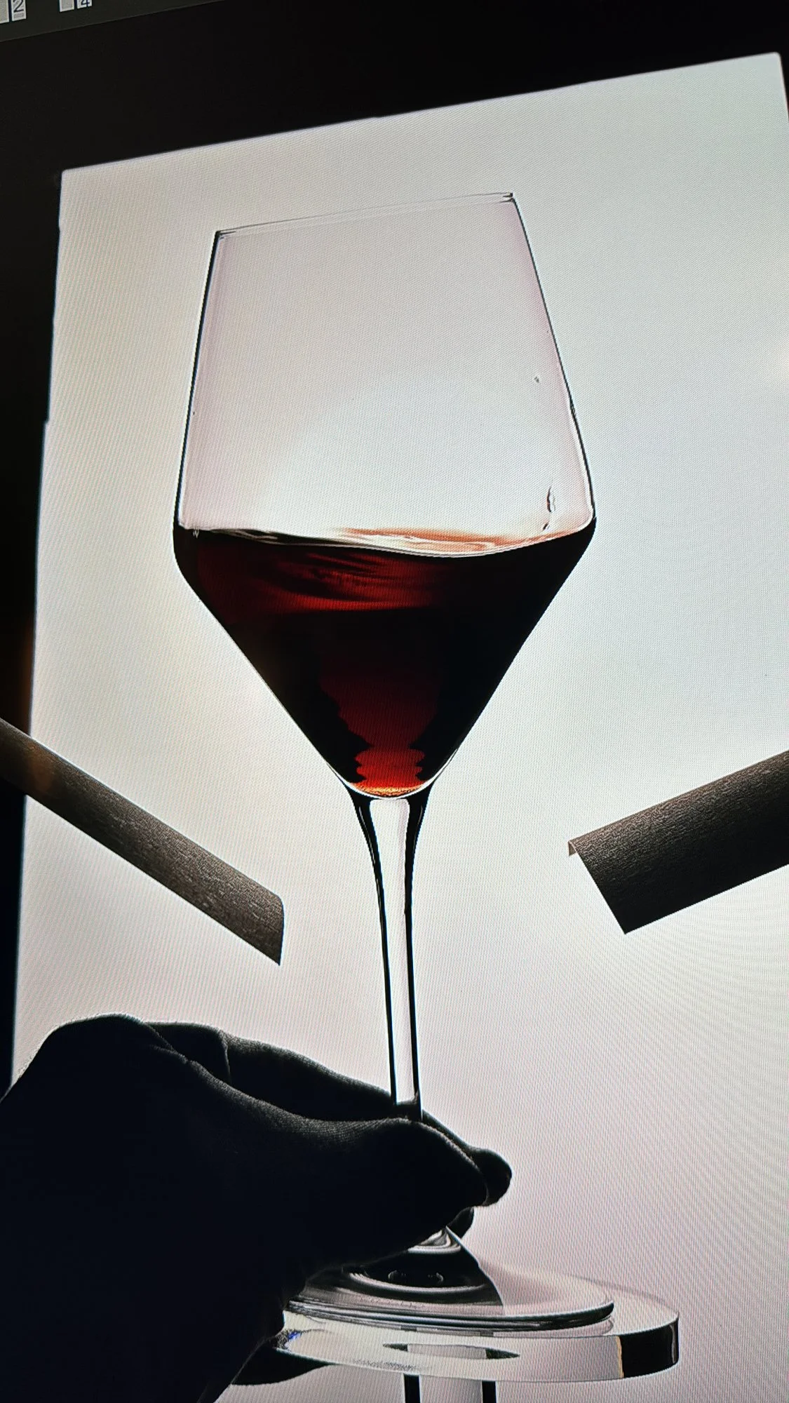 Ein Weinglas mit rotem Wein, das von einer Hand gehalten wird, vor einem hellen Hintergrund. Es gibt auch eine Hand, die das Glas stützt, und einige schwarze und graue Objekte im Hintergrund.