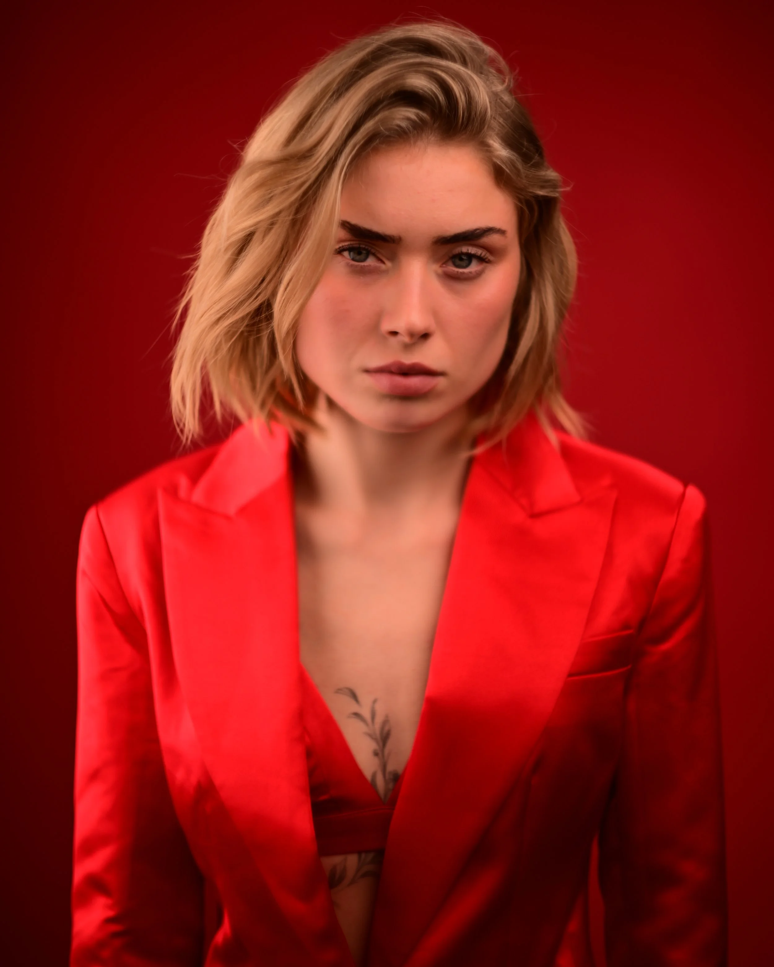 Porträt einer jungen Frau mit blonden, schulterlangen Haaren und blauen Augen, in einem roten Anzug, vor rotem Hintergrund, weist auf eine Tätowierung am Dekolleté hin.