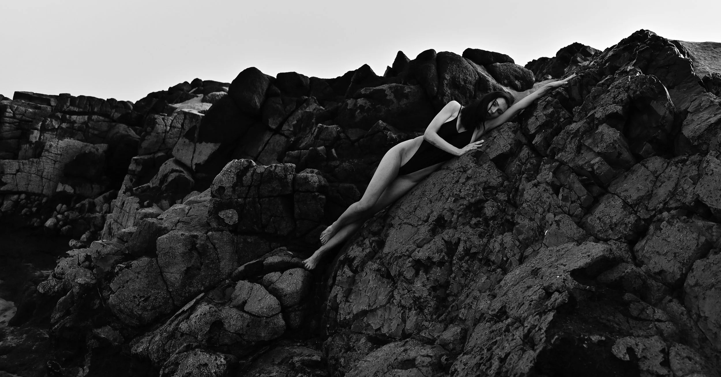 Schwarz-Weiß-Fotografie einer Frau in einem Badeanzug, die auf felsigem Küstengebiet liegt, mit Blickrichtung nach unten.