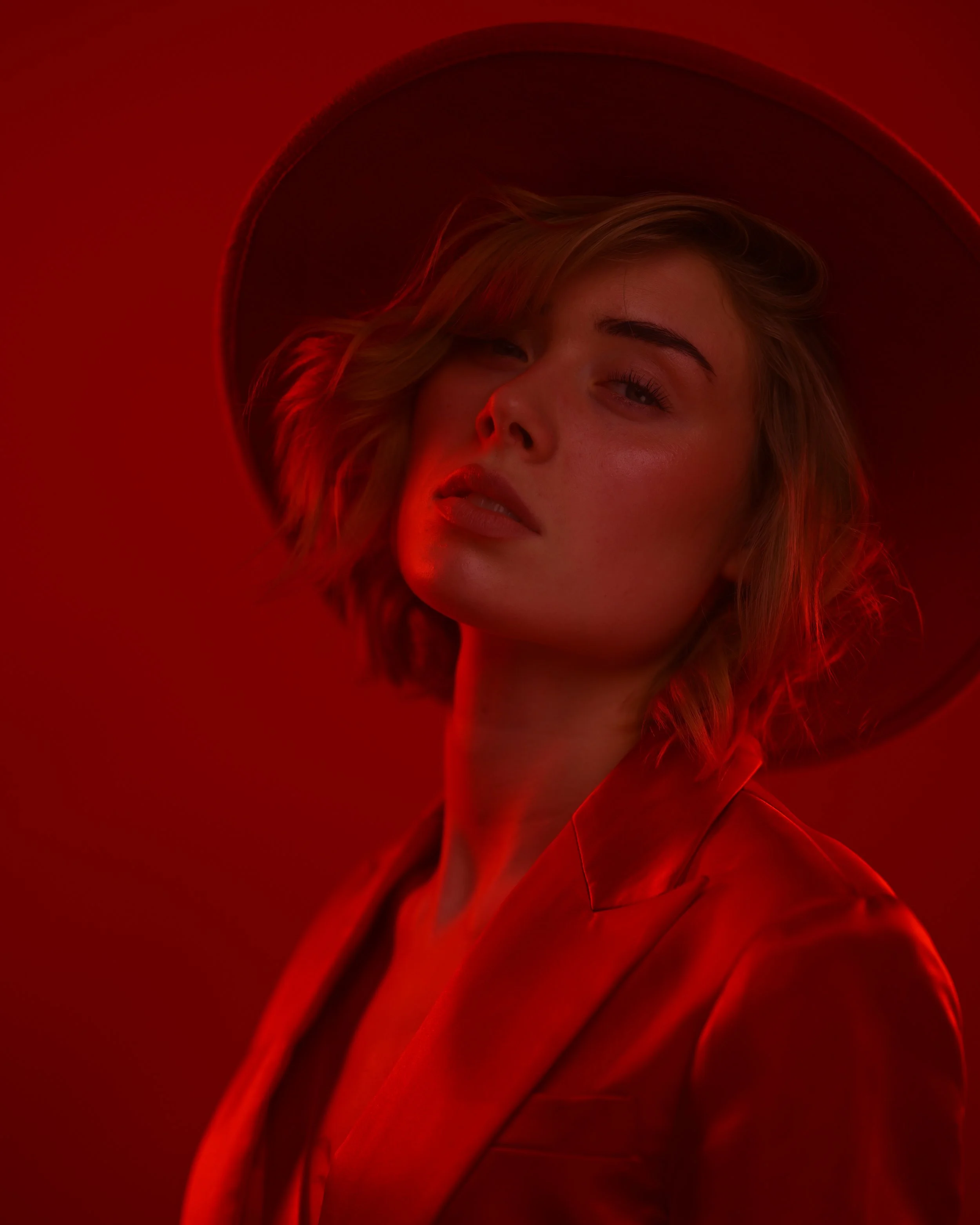 Porträt einer jungen Frau mit blondem Haar, die in einem roten Hut und rotem Mantel gegen einen roten Hintergrund posiert, Beleuchtung sorgt für dramatischen Effekt.