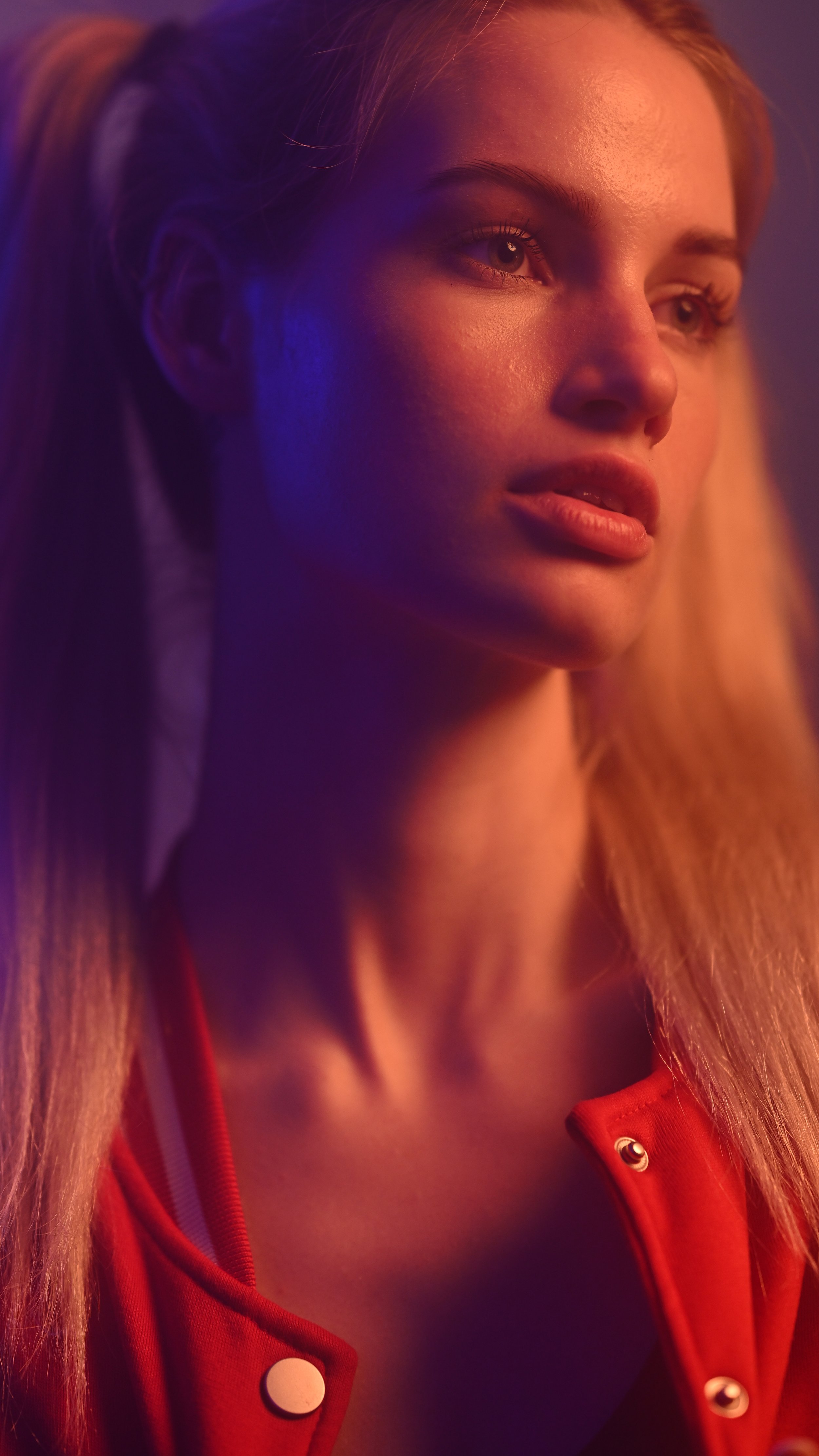 Nahaufnahme eines jungen Mädchens mit langen roten Haaren, das in warmes, farbiges Licht getaucht ist, trägt eine rote Jacke mit Knöpfen.