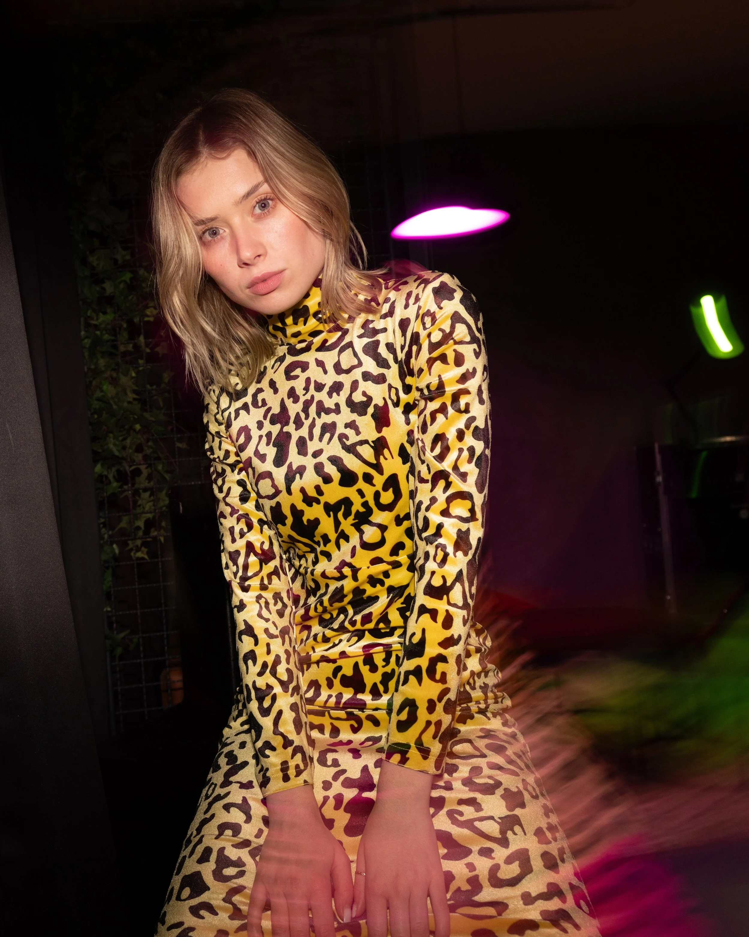 Ein junges Mädchen mit blonden Haaren, in einem gelben Leopardendruck-Kleid, sitzt in einer dunklen Umgebung mit farbigen Lichtakzenten.