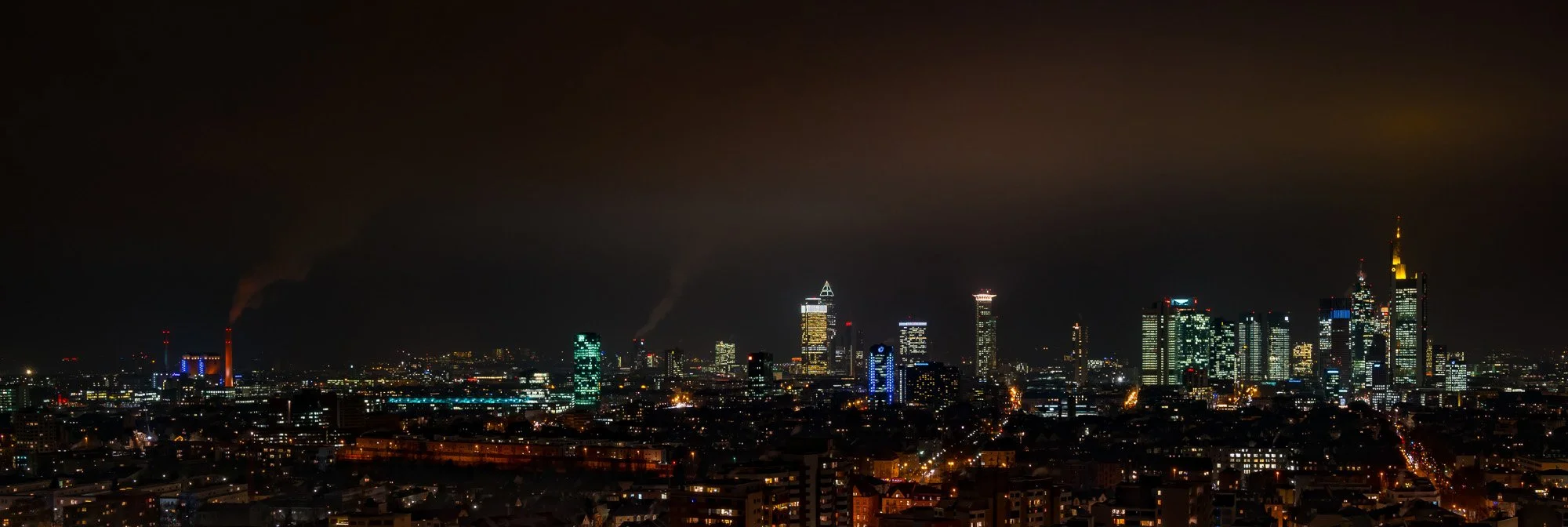 Nachthimmel über einer Stadt mit beleuchteten Hochhäusern und dunklen Rauchschwaden in der Ferne.