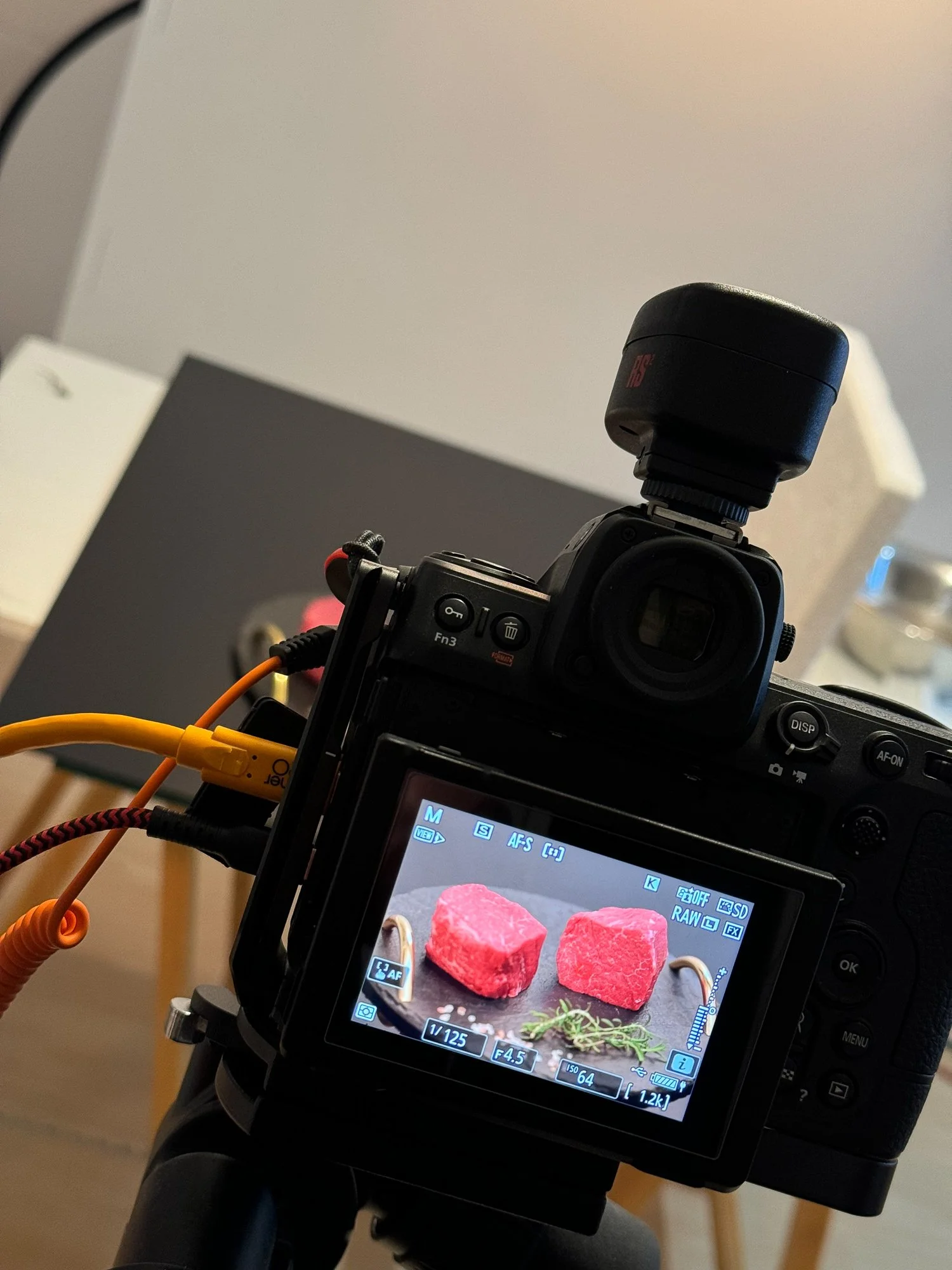 Eine digitale Kameraaufnahme zeigt vier rotes Fleischstücke auf einem schwarzen Grillplatte, mit einigen Kräutern, vor einem Display mit Kameraeinstellungen. Im Hintergrund sind unscharfe Einrichtungen.