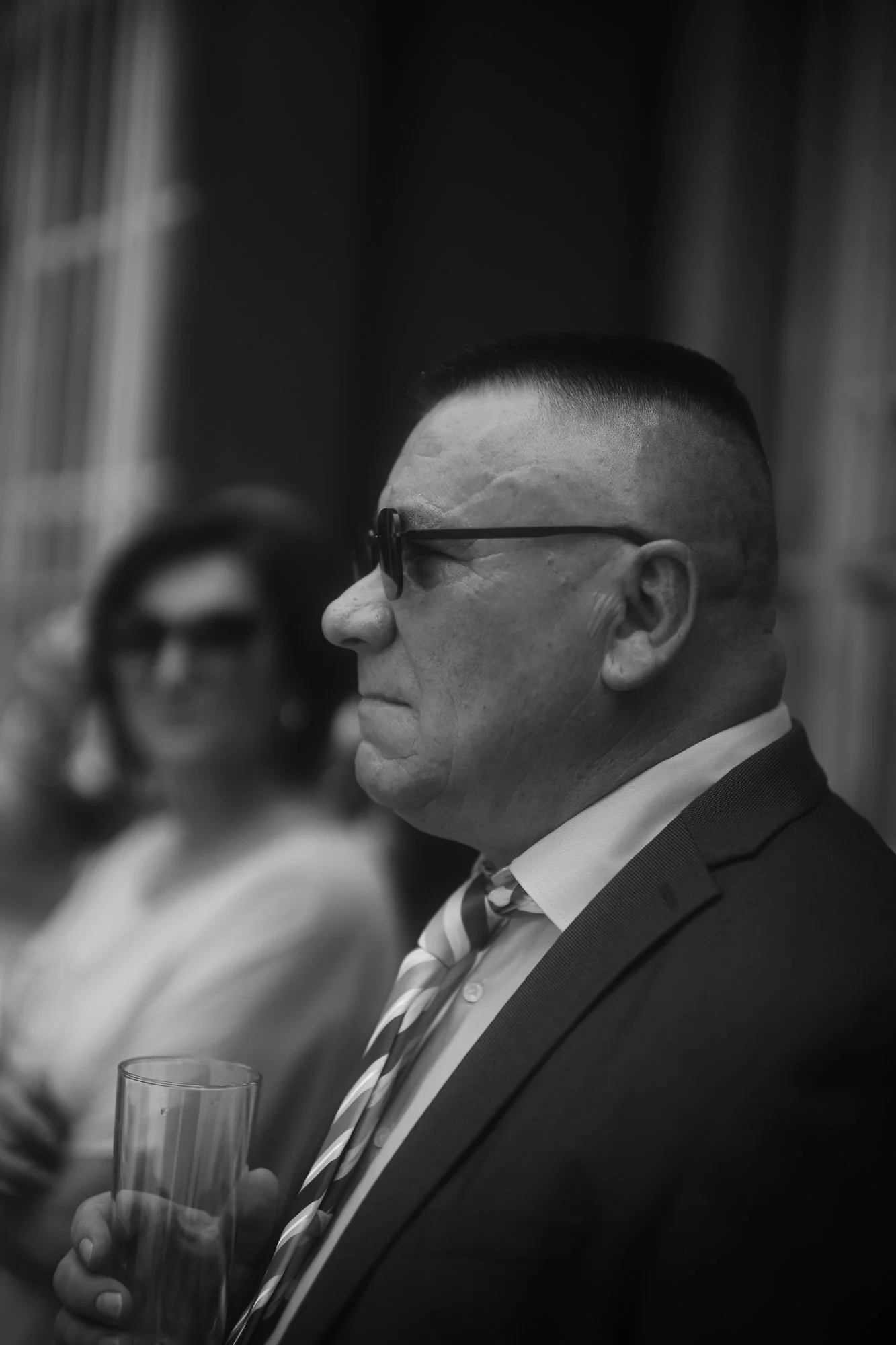 Schwarz-Weiß-Foto eines Mannes in Anzug und Krawatte, der eine Brille trägt und ein Glas in der Hand hält, bei einer formellen Veranstaltung.