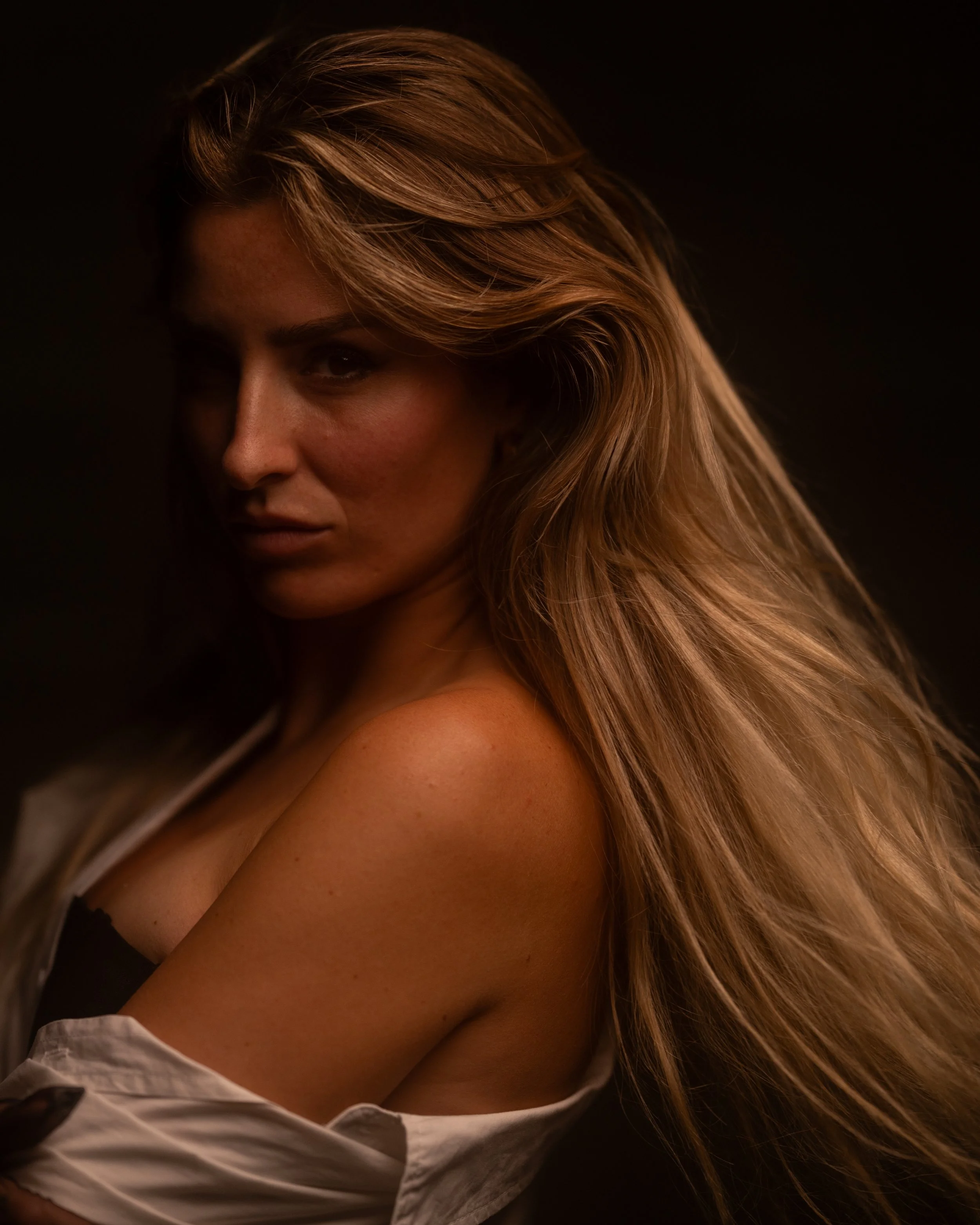 Nahaufnahme eines Teils eines Frauenkopfes mit langem, lockigem, braunem Haar. Das Gesicht ist nur teilweise sichtbar, vor einem dunklen Hintergrund.