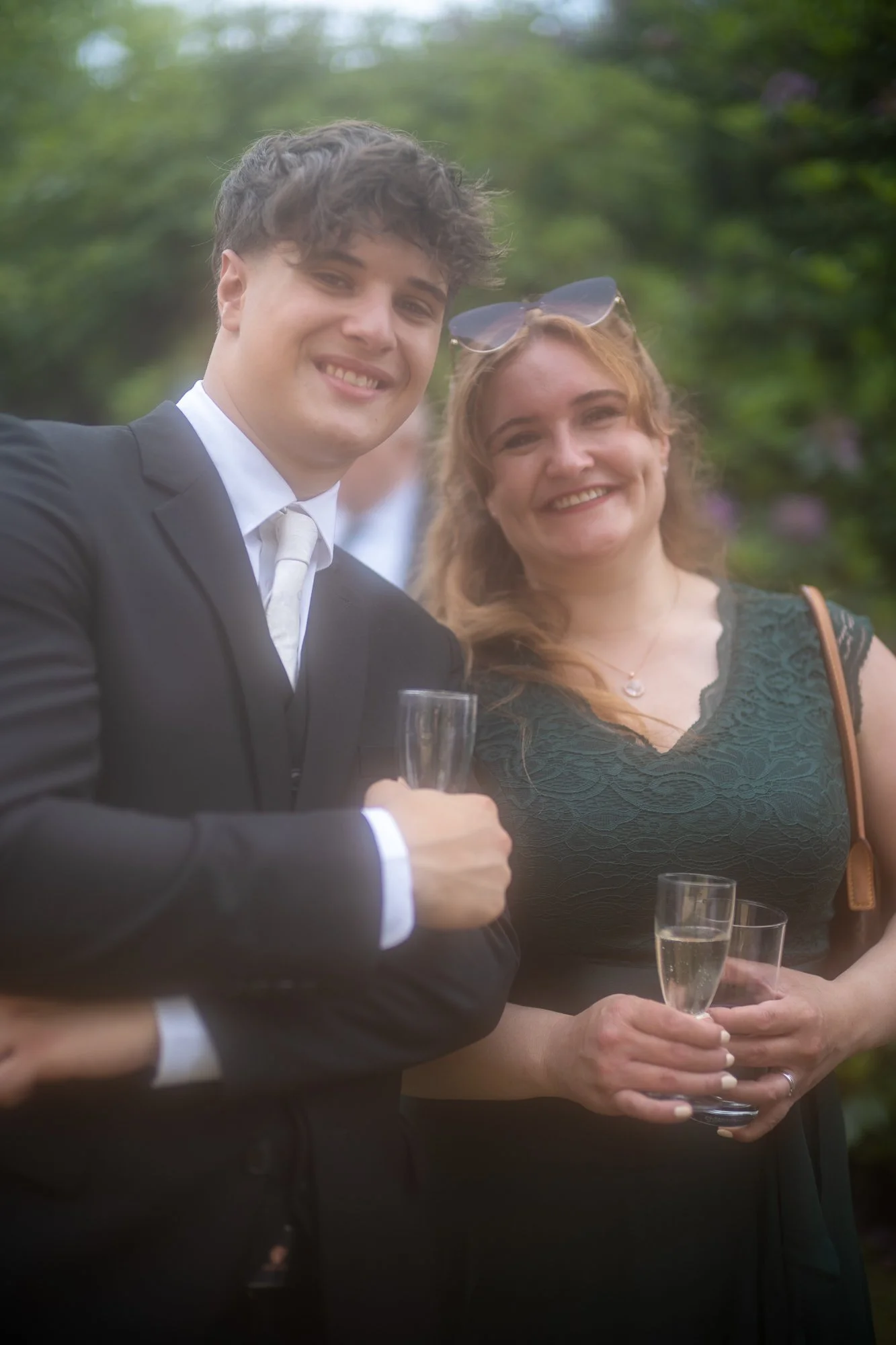Zwei lachende Menschen, ein Mann im Anzug und eine Frau in einem grünen Kleid, halten Champagnergläser auf einer Feier im Freien, umgeben von grünen Bäumen.