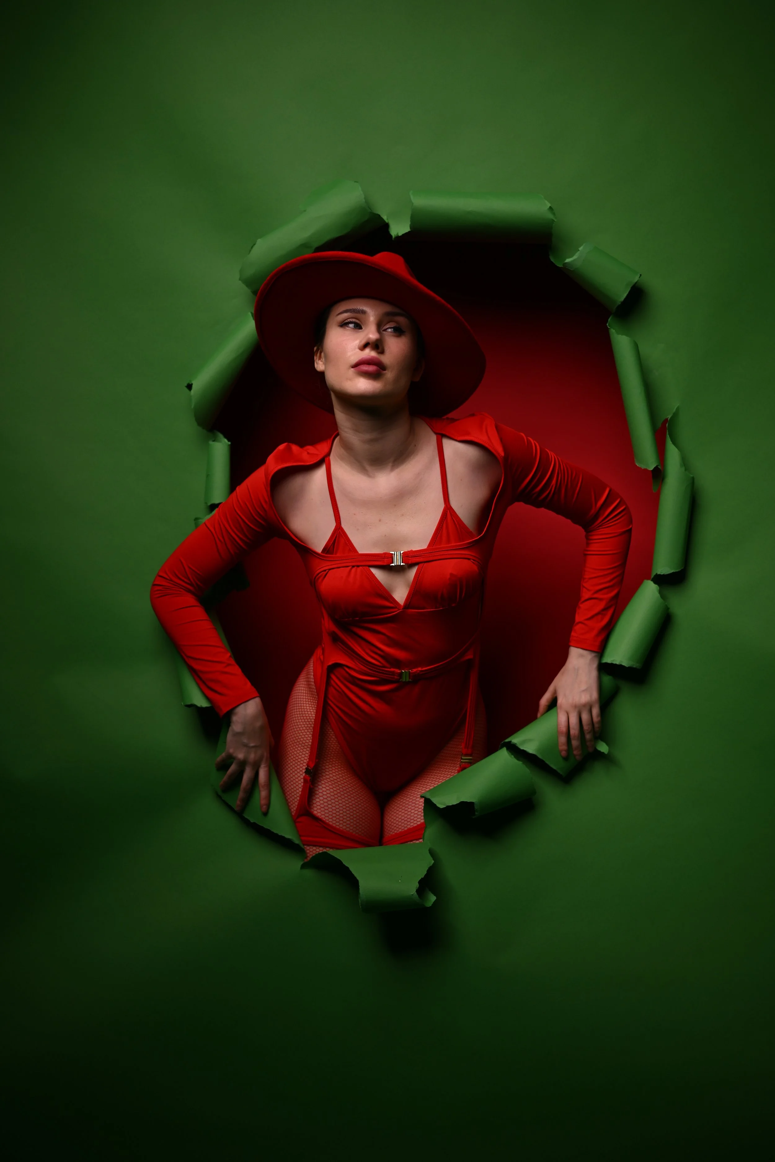 Frau in rotem Kleid und Hut, die aus einer grün ummantelten Öffnung schaut.