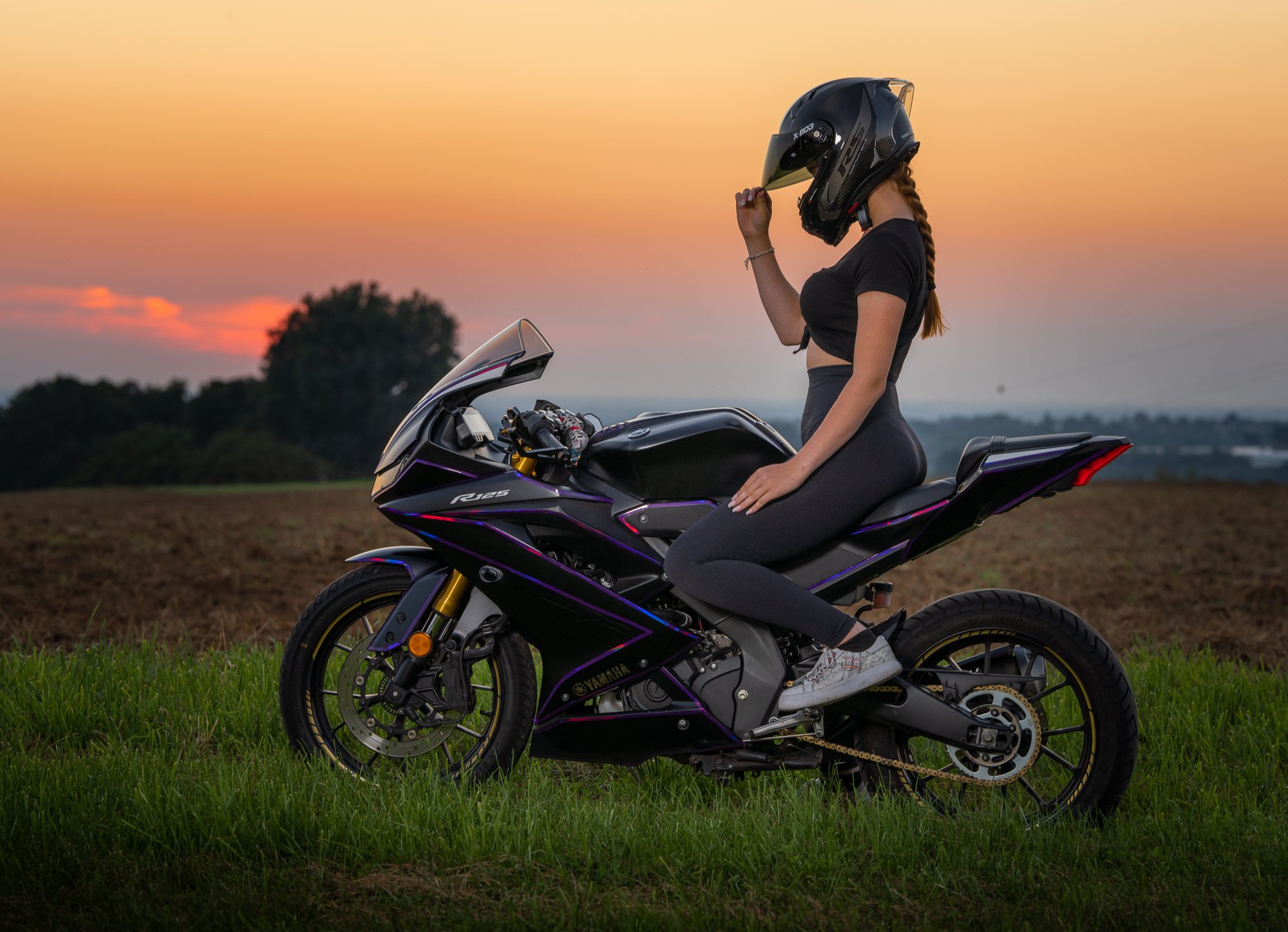 Eine Frau sitzt auf einer schwarzen Yamaha R125 Motorrad im Grünen bei Sonnenuntergang, trägt einen schwarzen Helm, ein schwarzes T-Shirt und schwarze Leggings.
