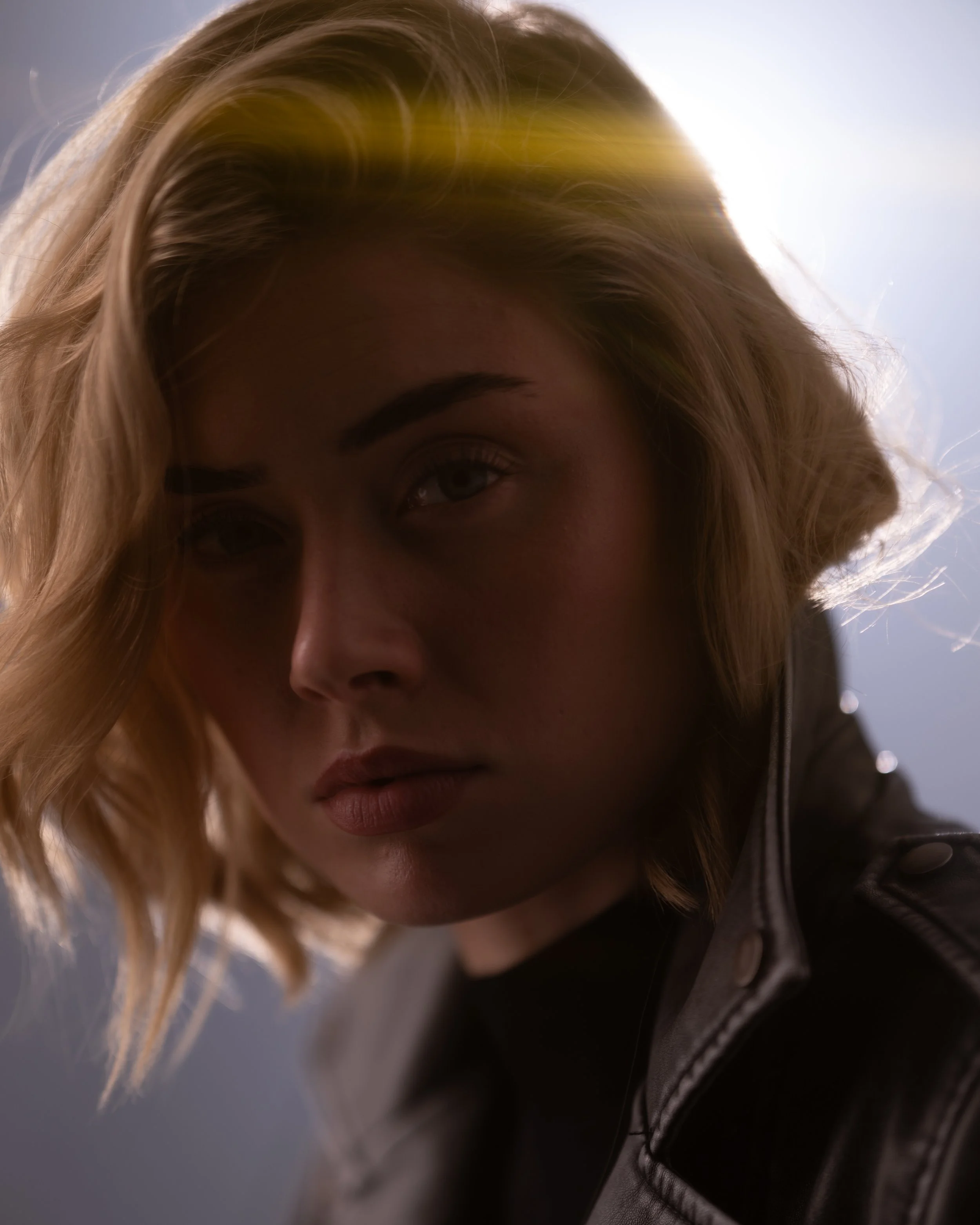 Nahaufnahme eines Gesichts einer jungen Frau mit blonden, lockigen Haaren, die leicht über die Schulter fallen, und dunklem Oberteil, vor hellem Hintergrund.