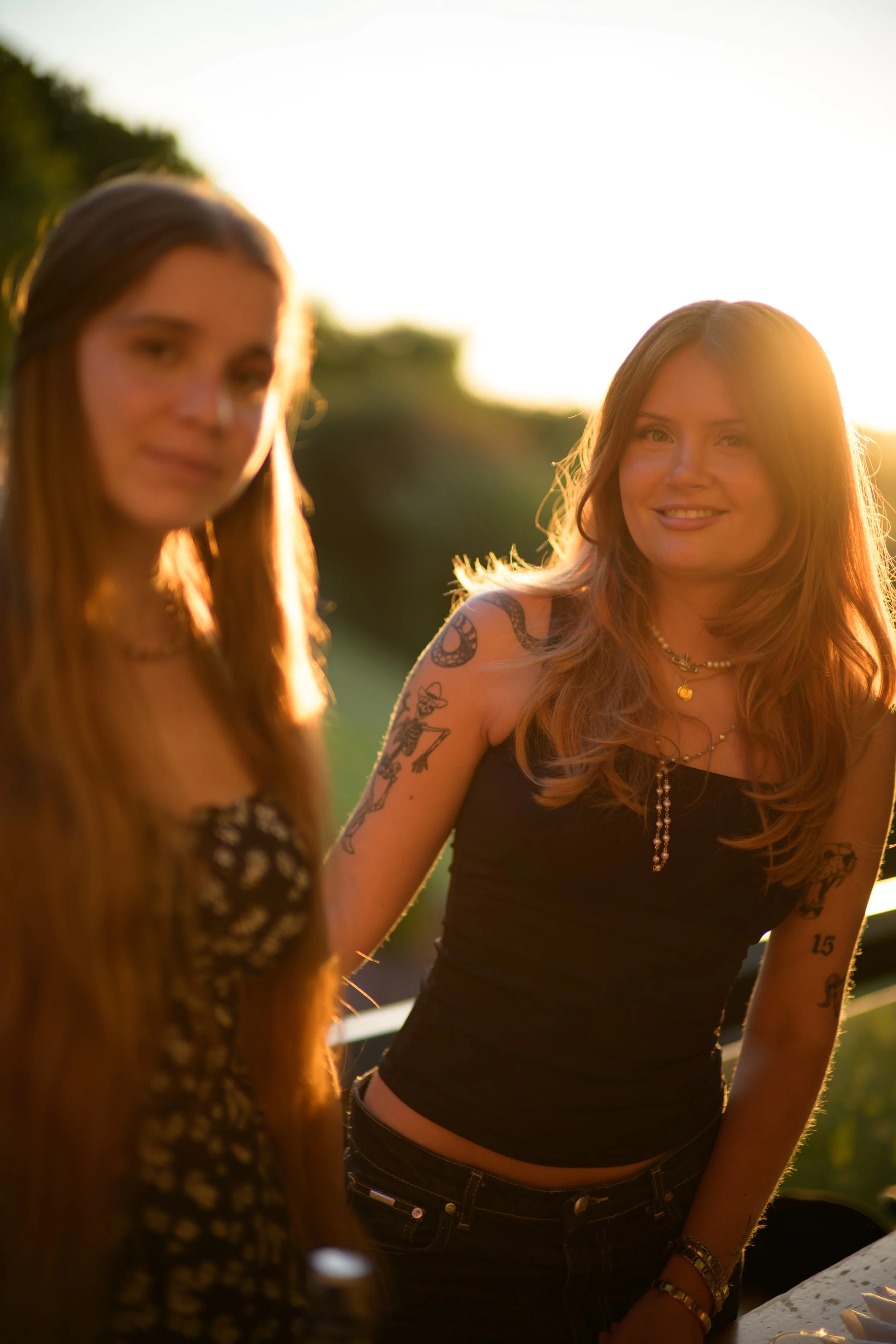 Zwei Frauen mit langen Haaren, tätowiert, bei Sonnenuntergang im Freien, lächelnd, in einem lockeren Gespräch.