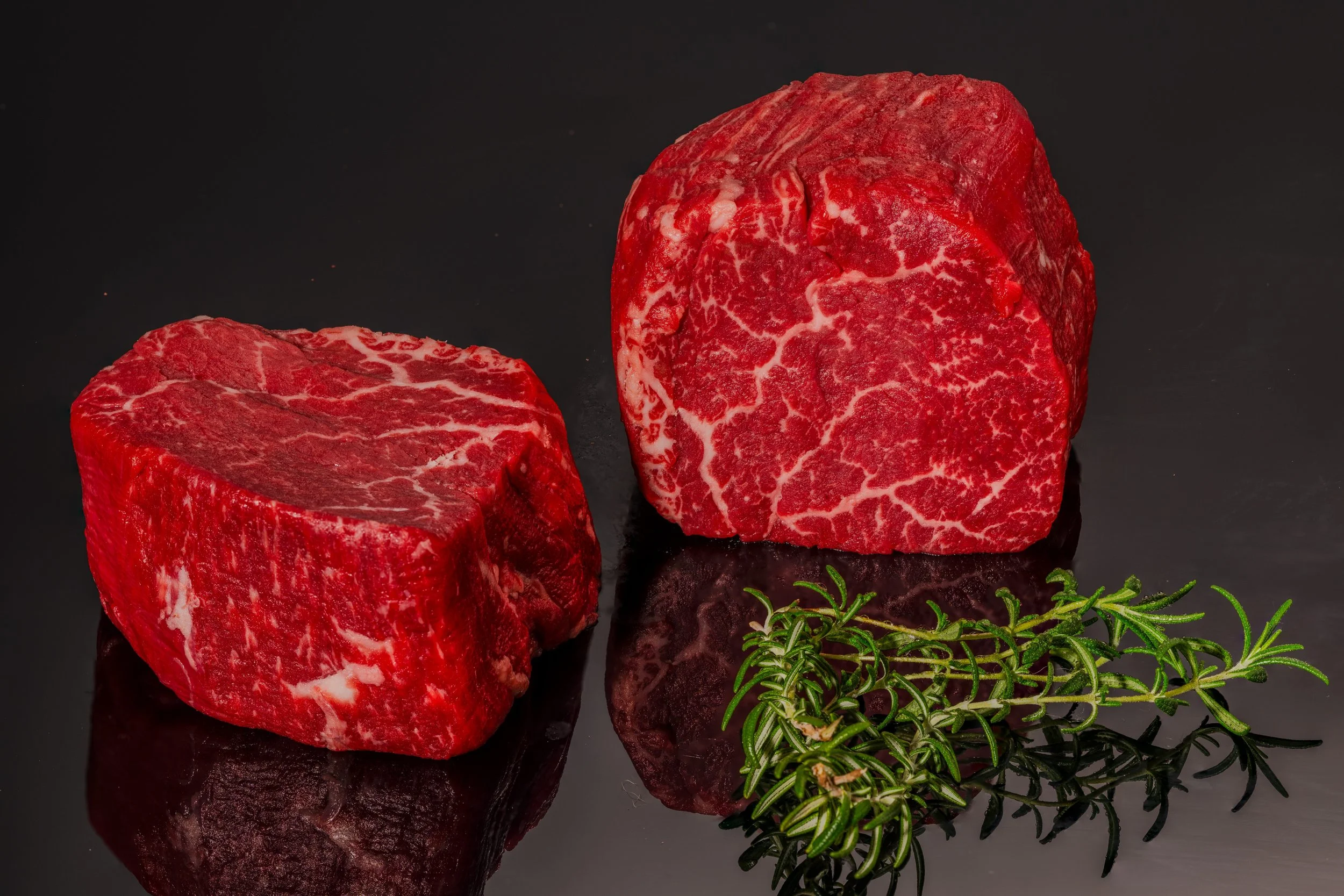 Zwei Stücke Rindfleisch, eines aufrecht stehend, eines liegend, neben grünen Kräutern auf einem schwarzen spiegelnden Hintergrund.