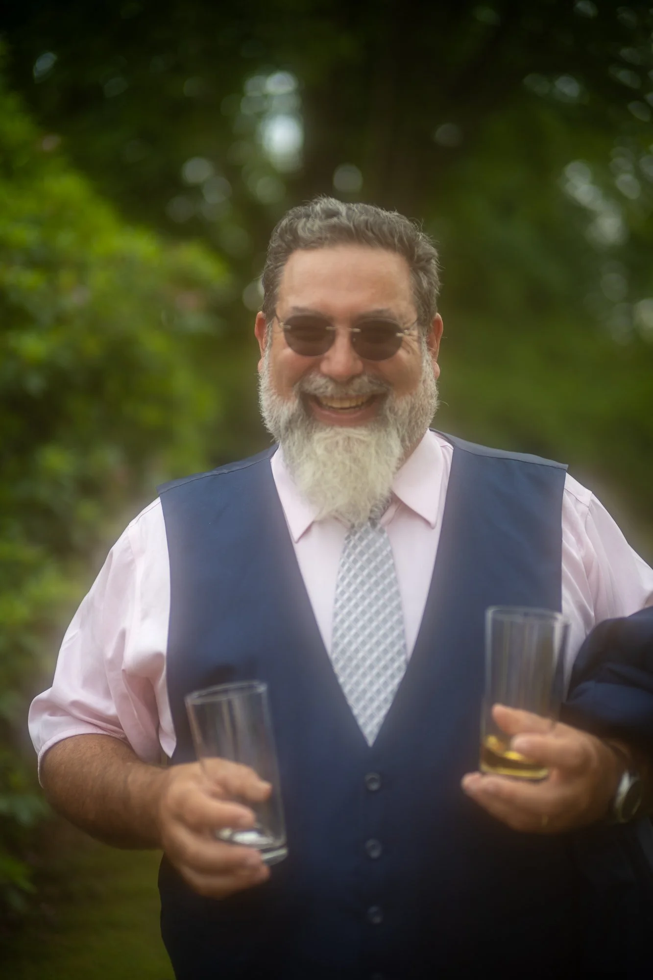 Ein lachender Mann mit Brille, weißem Bart und grauen Haaren trägt ein weißes Hemd, eine dunkelblaue Weste und eine Krawatte. Er hält zwei Gläser mit einem Getränk in den Händen. Der Hintergrund ist grün und bewaldet.
