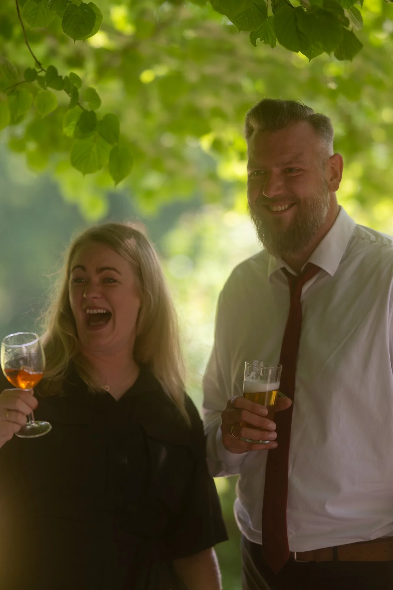 Zwei Menschen, eine Frau und ein Mann, lachen und halten Getränke in einem schönen, grünen, outdoor Umfeld bei einer Feier.