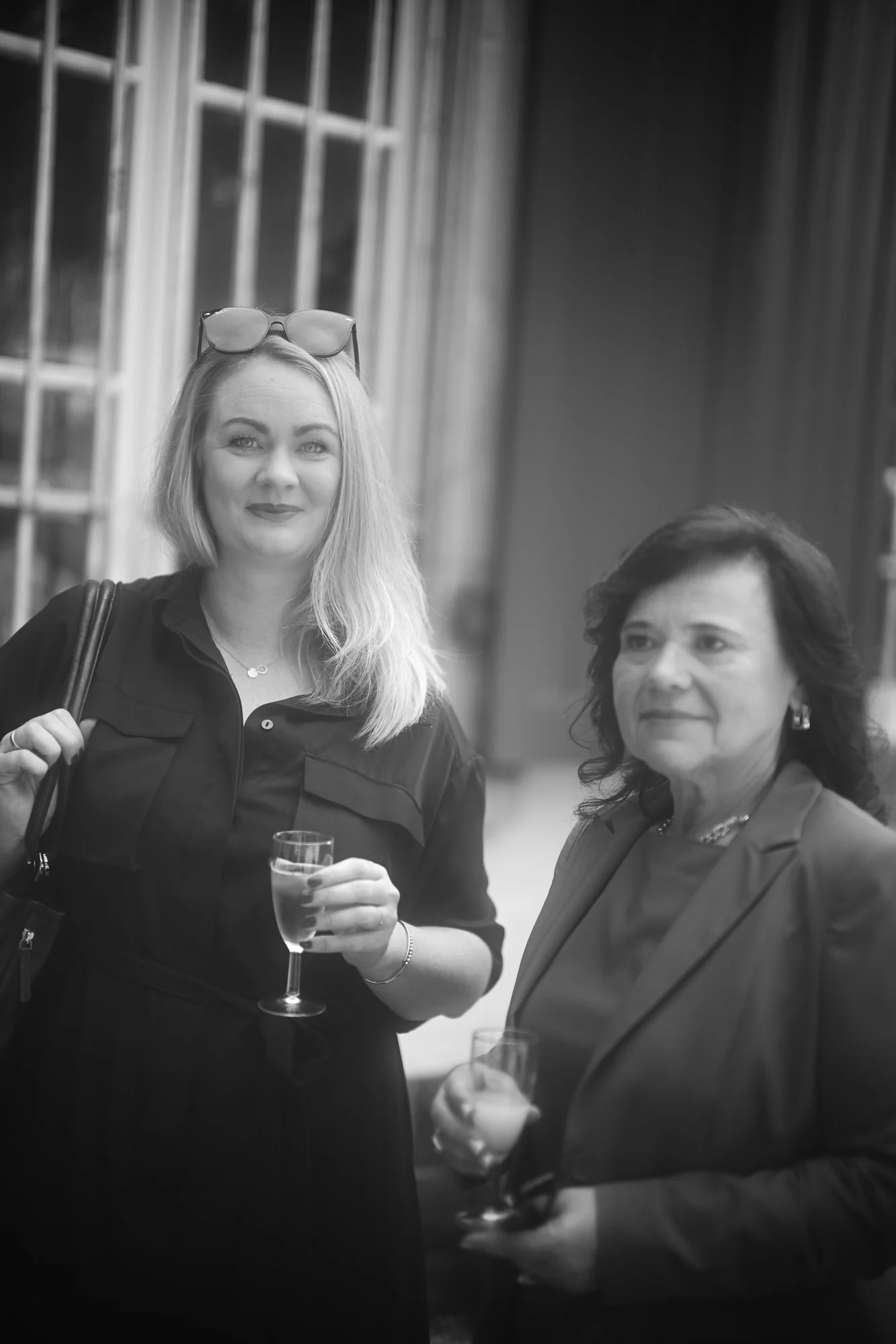 Zwei Frauen bei einer socialen Veranstaltung, eine hält ein Glas Sekt, im Hintergrund ein Fenster mit Gittern.