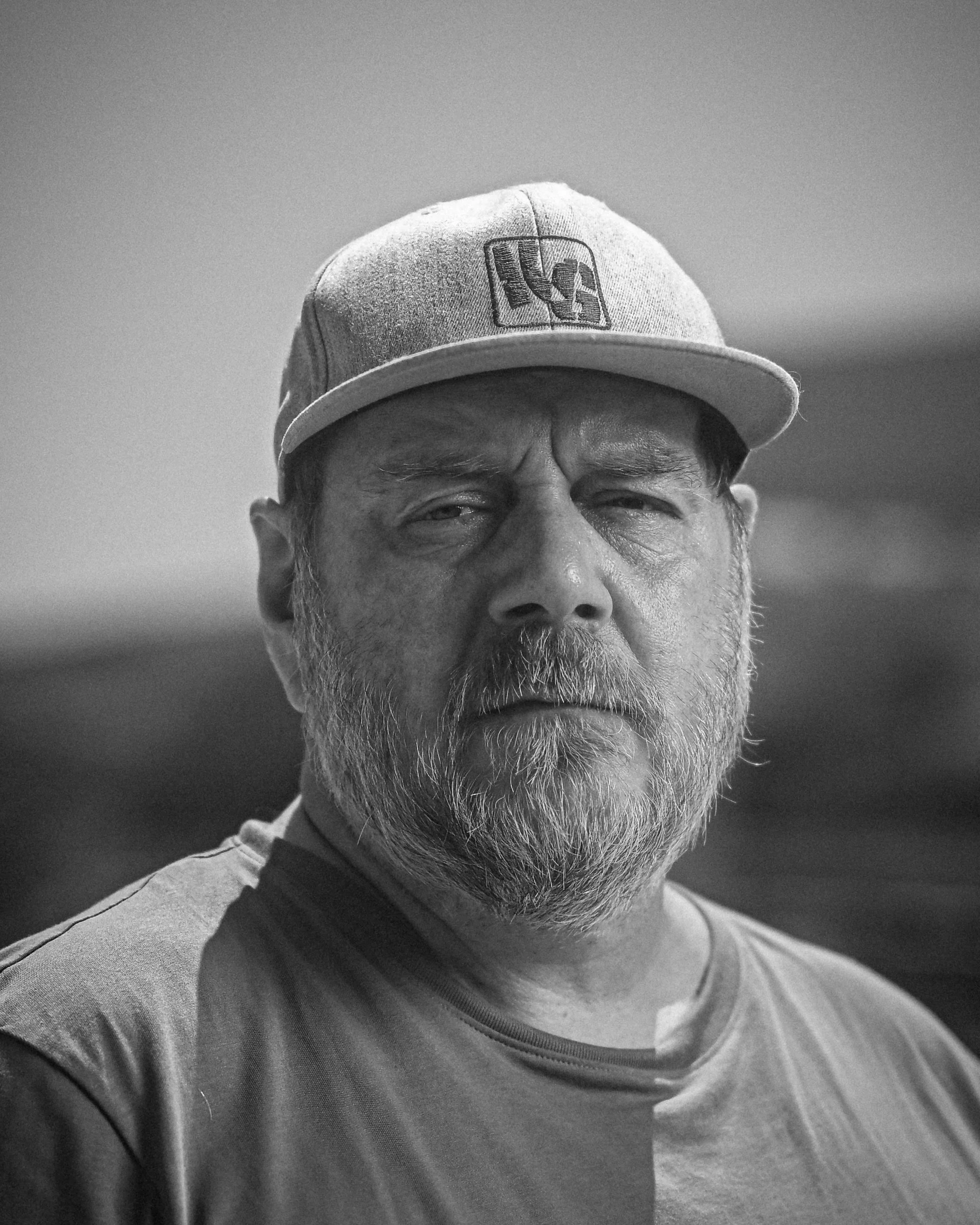 Ein älterer Mann trägt eine Baseballmütze mit einem Logo und eine graue T-Shirt, er hat einen dichten Bart und einen ernsten Gesichtsausdruck.