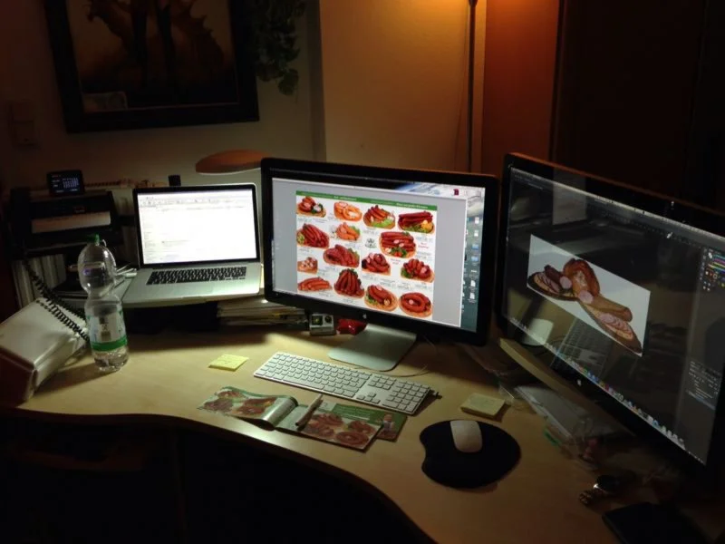 Arbeitsplatz mit zwei Monitoren, einem Laptop, Tastatur, Maus, Wasserflasche und Skizzen, die Essensbilder zeigen.