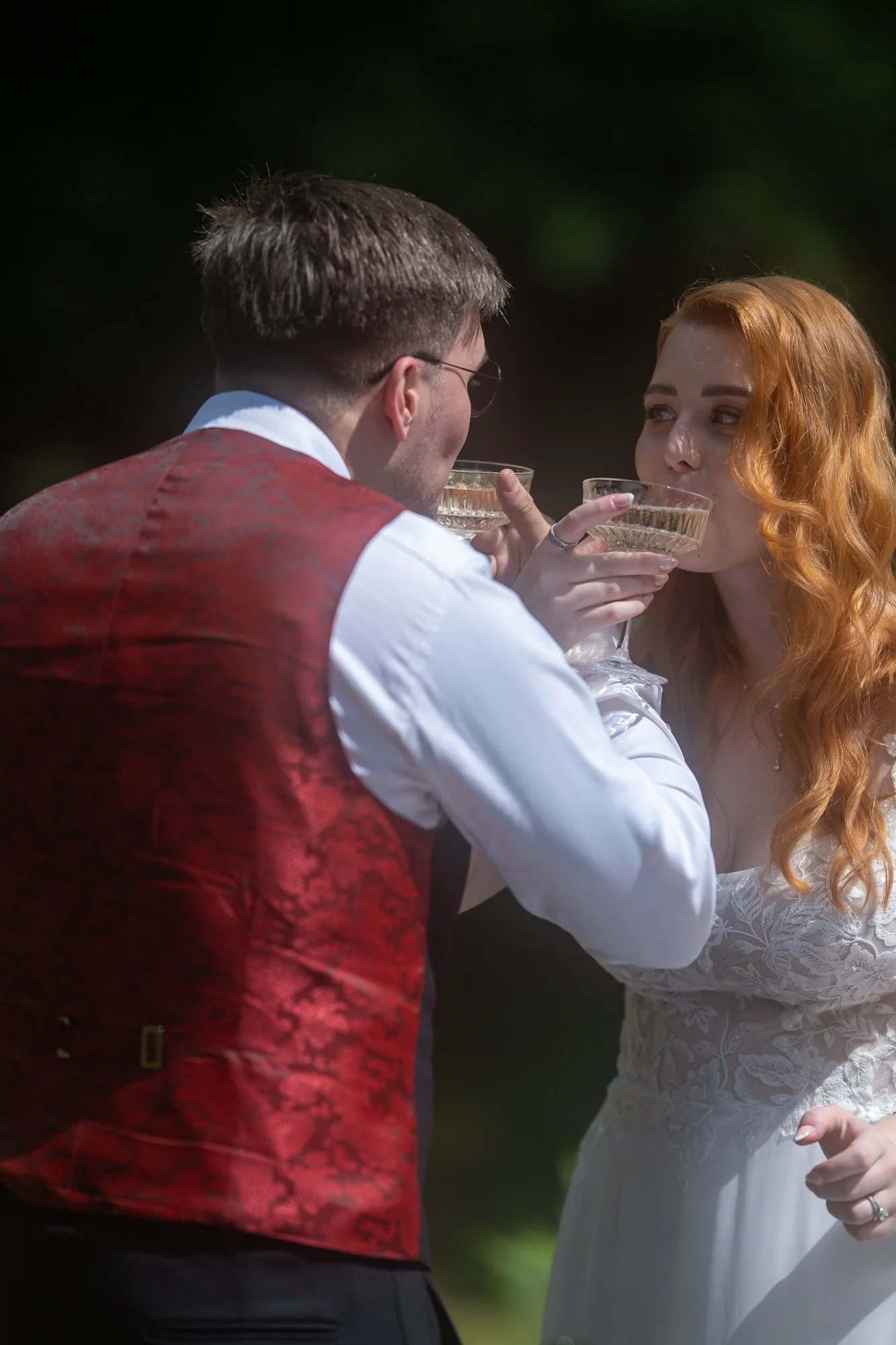 Ein Mann und eine Frau trinken gemeinsam aus Kristallgläsern. Die Frau trägt ein weißes Kleid mit Spitze, der Mann trägt ein weißes Hemd und eine rote Weste.