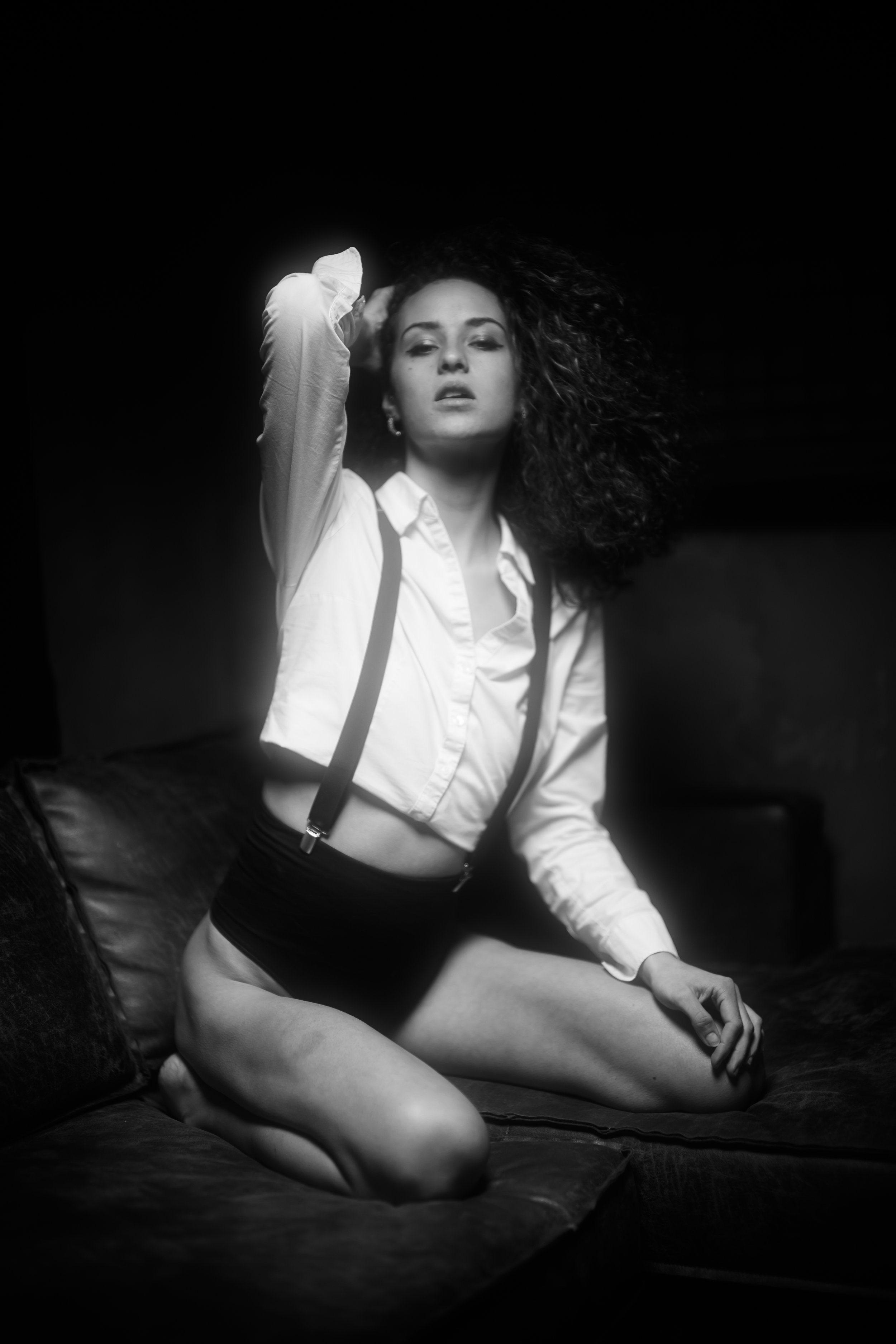 Schwarze und weiße Fotografie eines sexy Frauenporträts. Frau trägt eine weiße Hemdbluse, schwarze Shorts und Hosenträger, sitzt auf einem Bett mit einem schwarzen Kopfteil, haar- und gestischer Ausdruck, mit einem selbstbewussten Blick.