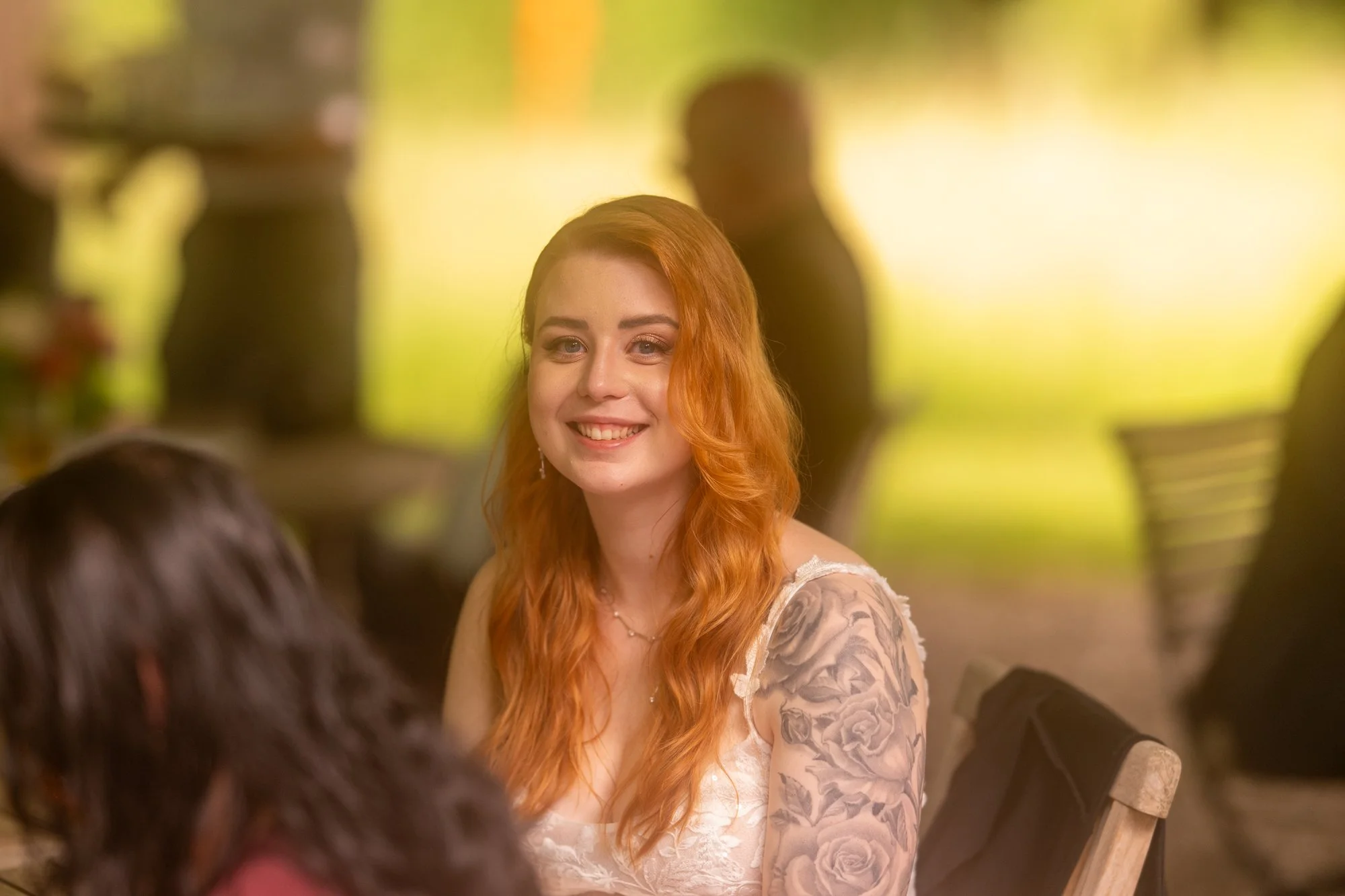 Eine lachende junge Frau mit roten Haaren und Tätowierungen auf dem Arm, im Gespräch mit einem Mann, im Hintergrund verschwommene Silhouetten anderer Personen und gelbe Beleuchtung.