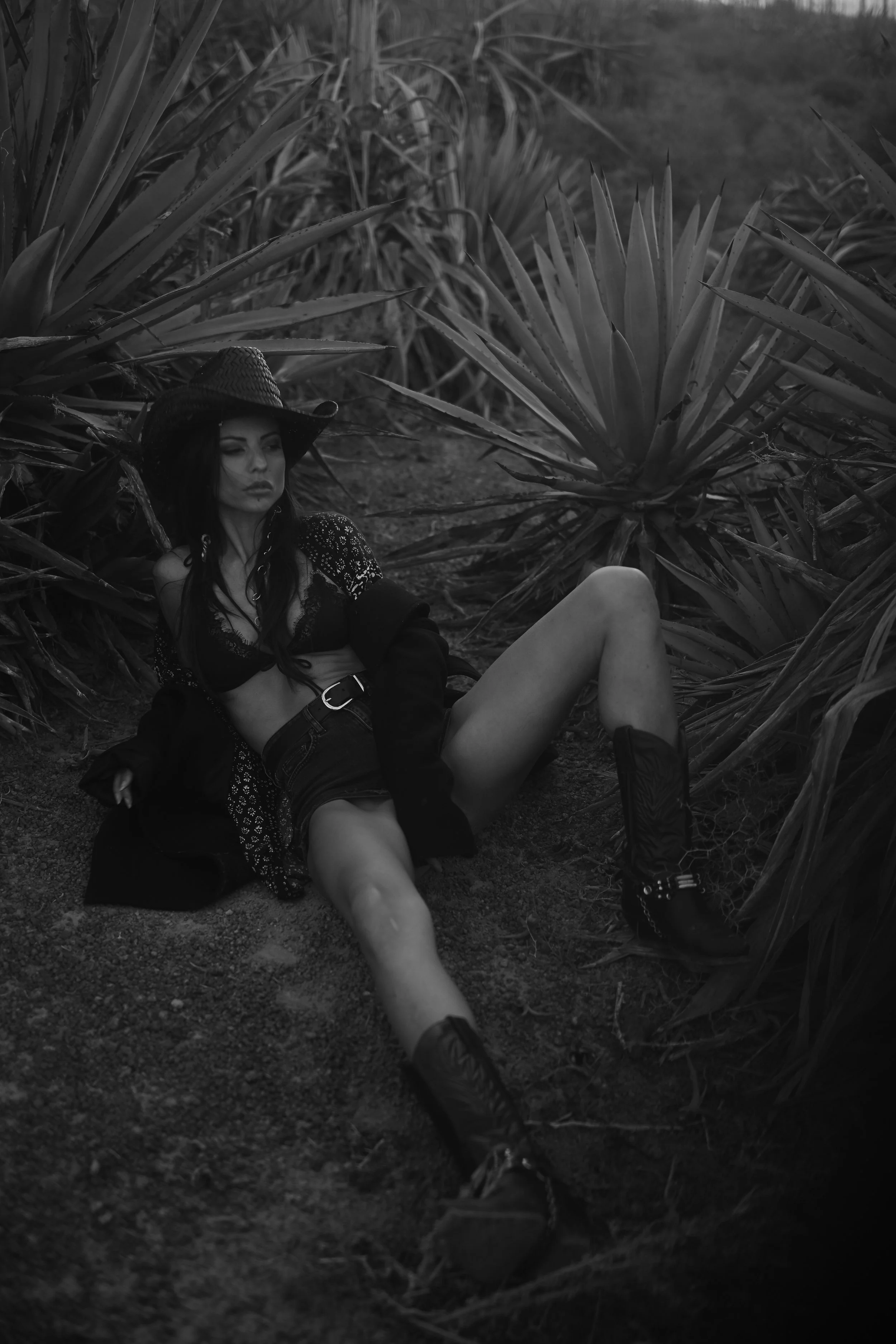 Eine Frau liegt im Boden in einem Garten mit großen Pflanzen und trägt einen Hut, eine transparente Bluse, Shorts und Stiefel.