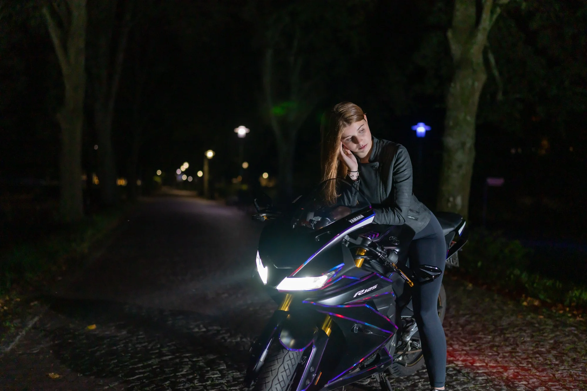 Ein junges Mädchen sitzt auf einem schwarzen Motorrad in einer dunklen, nächtlichen Umgebung mit Bäumen. Es trägt eine schwarze Jacke und sieht nachdenklich aus.